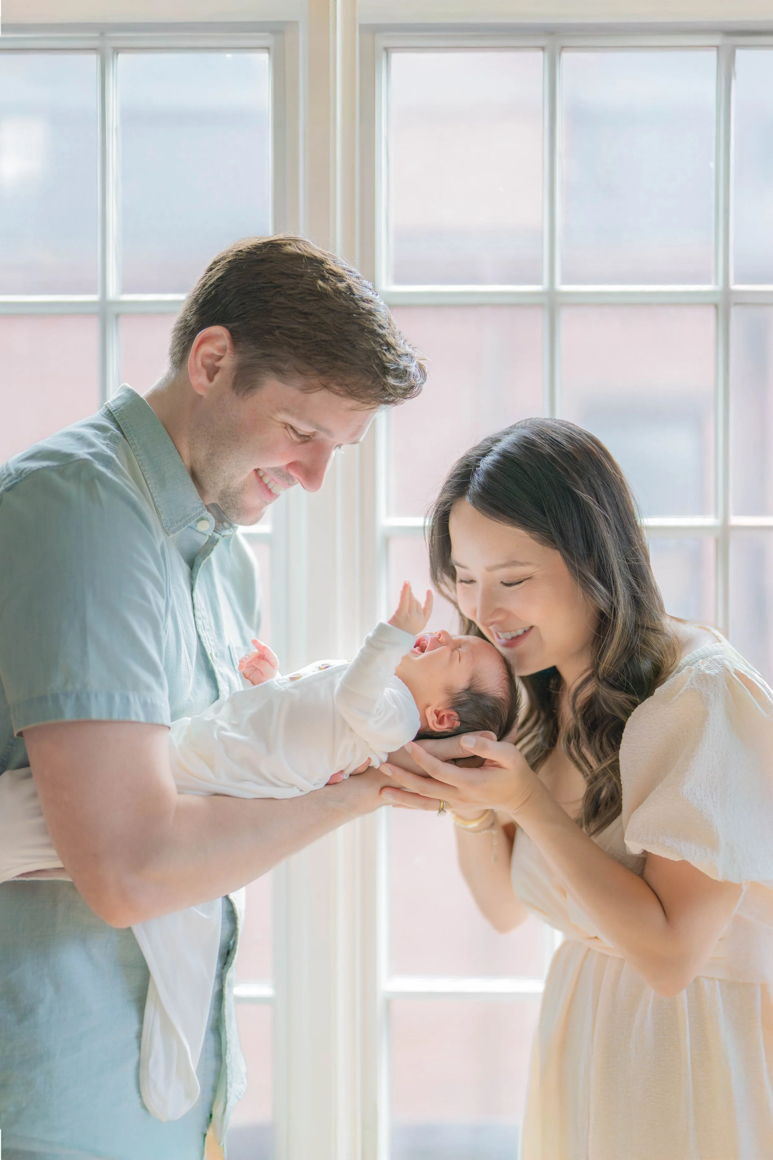 A joyful young couple holding a newborn baby, standing by a large window with grid panes in a bright room.