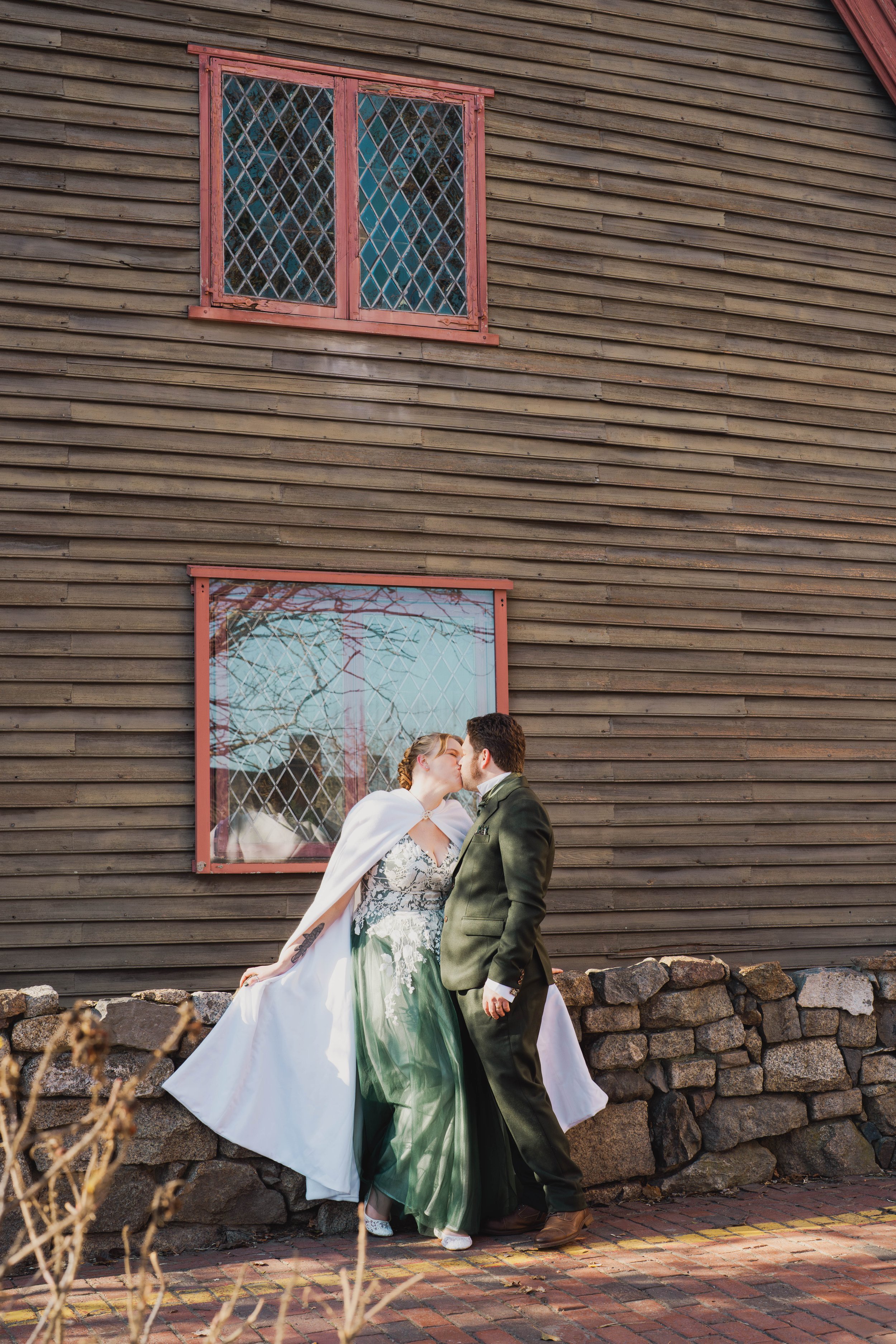 A couple dressed in wedding attire sharing a kiss outside a wooden building with two decorated windows, with rocks and dried plants in the foreground.