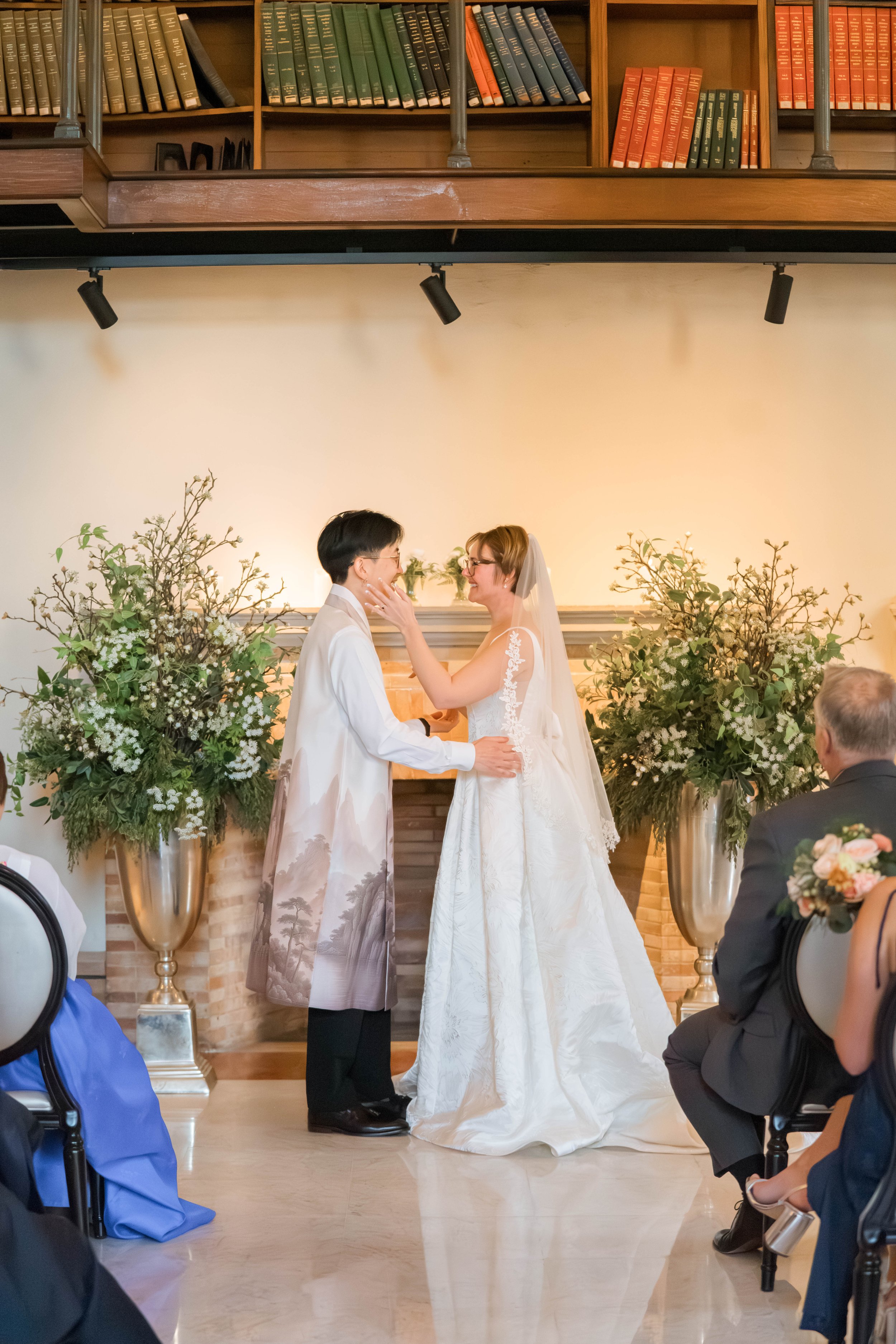 A wedding ceremony with two women, one in a white wedding dress with a veil and the other in black and white attire, exchanging vows. They are standing between large floral arrangements with green foliage and white flowers, in a room with a wooden sh