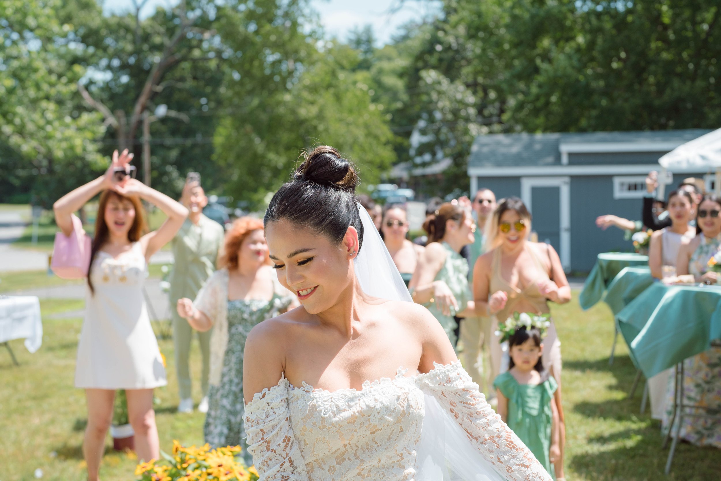 A bride in a white off-shoulder lace wedding dress smiling as she walks outdoors on a sunny day. Behind her, a group of women and girls, some in floral or pastel dresses, are gathered in a lush green yard, celebrating and taking photos. There are tre
