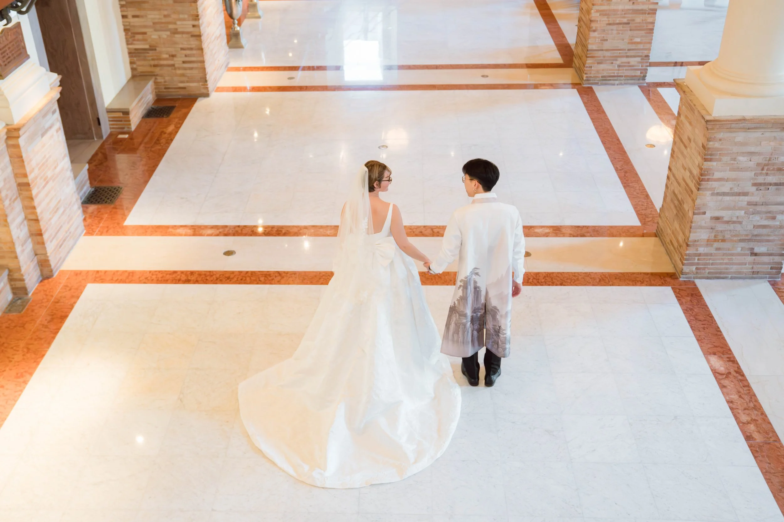 A bride and groom holding hands inside a spacious, ornate building with polished marble floors and brick columns.