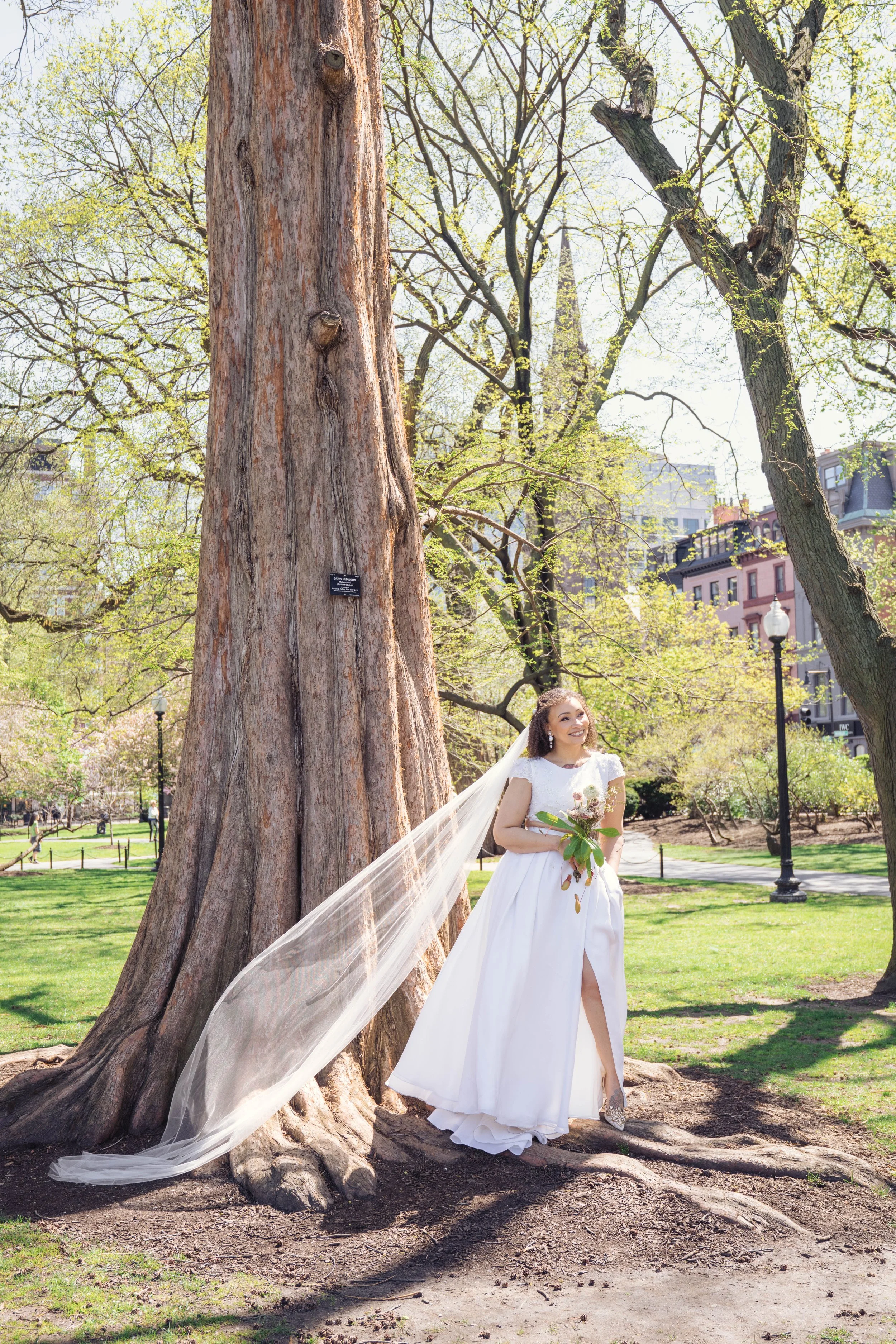A bride in a white wedding gown with a veil, holding a bouquet of flowers, standing next to a large tree in a park on a sunny day. She is smiling and looking towards the camera, with a cityscape and buildings visible in the background.