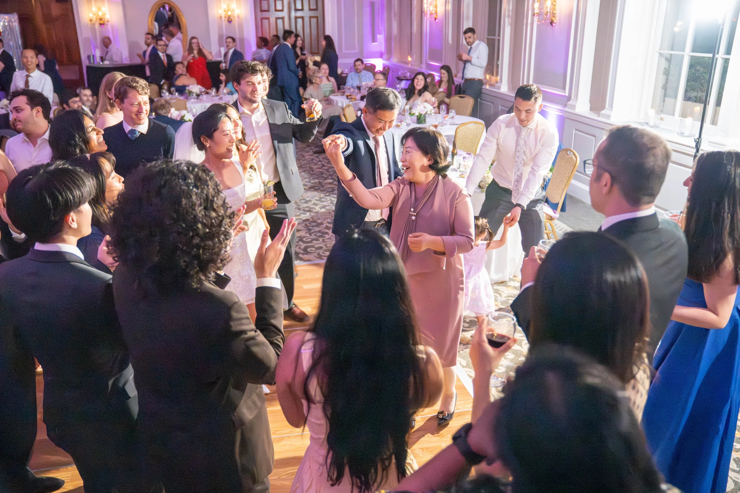 People dancing and socializing at a lively party or celebration in an elegant decorated room with large windows and purple lighting.