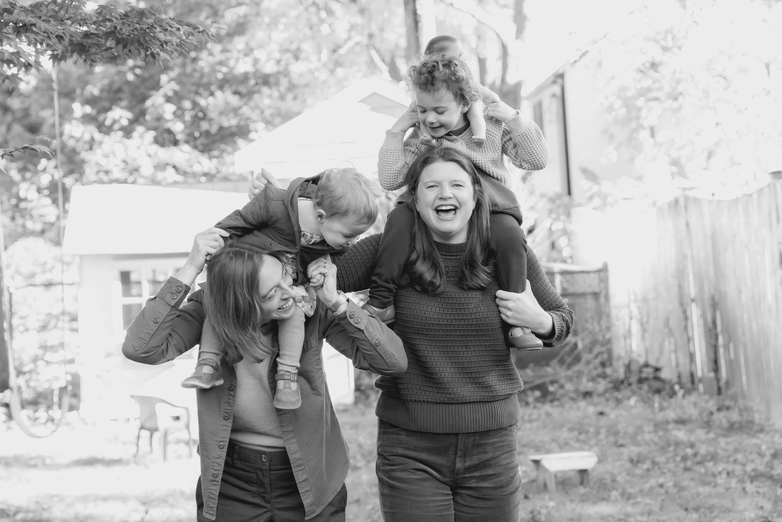 A woman is playing with four kids outdoors, with one kid on her shoulders, in a backyard with trees and a house in the background.