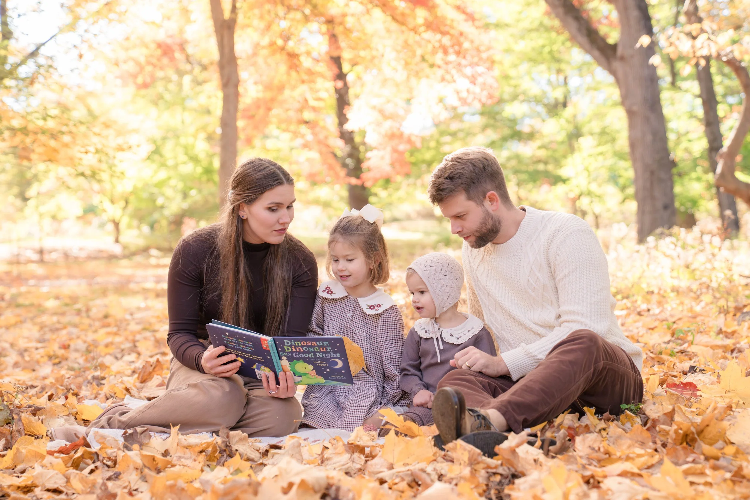 A family of four, including two small children, sits on the ground among fallen autumn leaves in a park, looking at a children's book titled "Dinosaur Dinosaur Say Good Night".