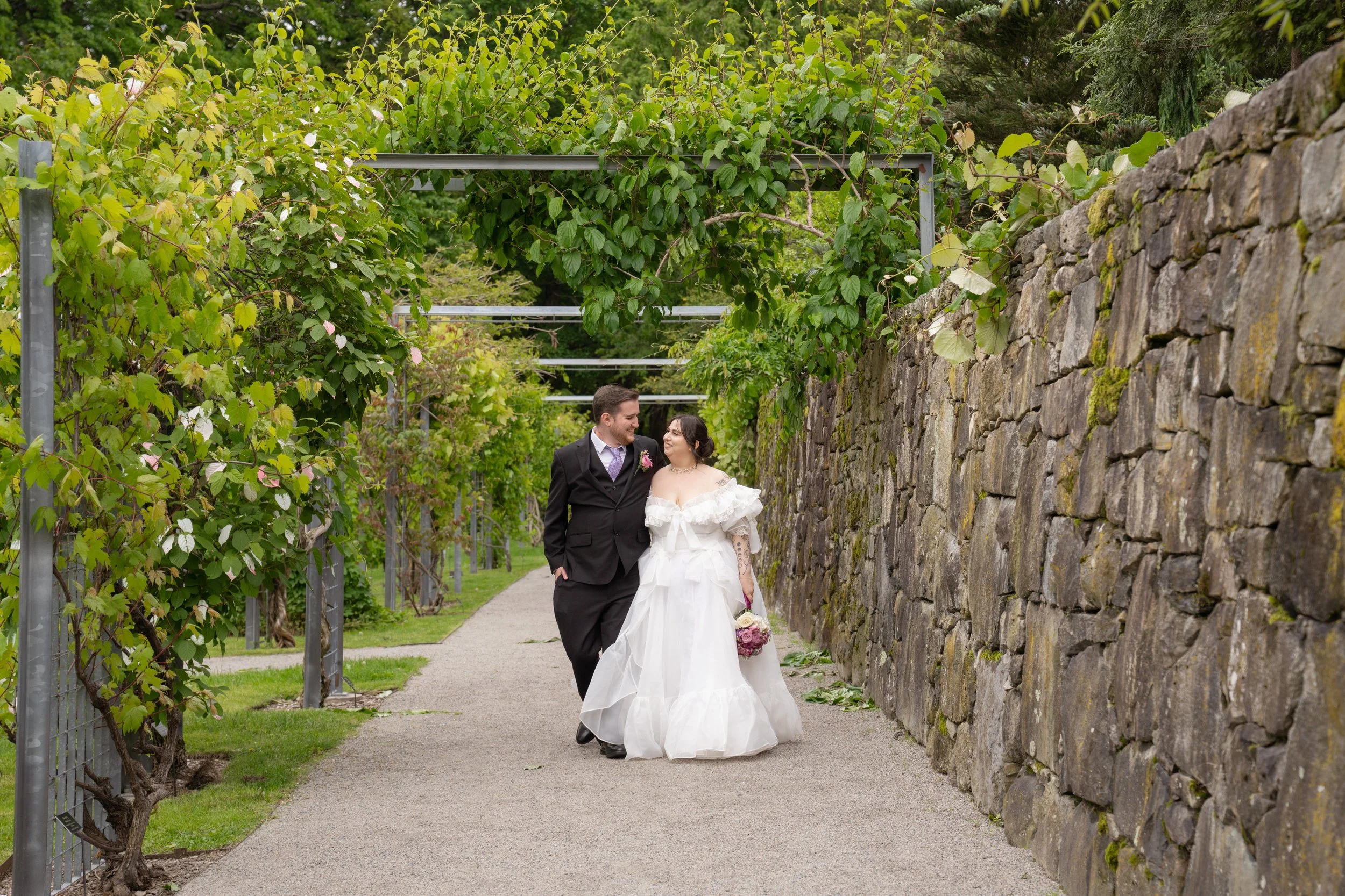 A bride and groom walking together in an outdoor garden setting, smiling at each other. The groom is dressed in a black suit with a purple tie, and the bride is in a white wedding gown holding a bouquet. They are walking beside a stone wall and a pat