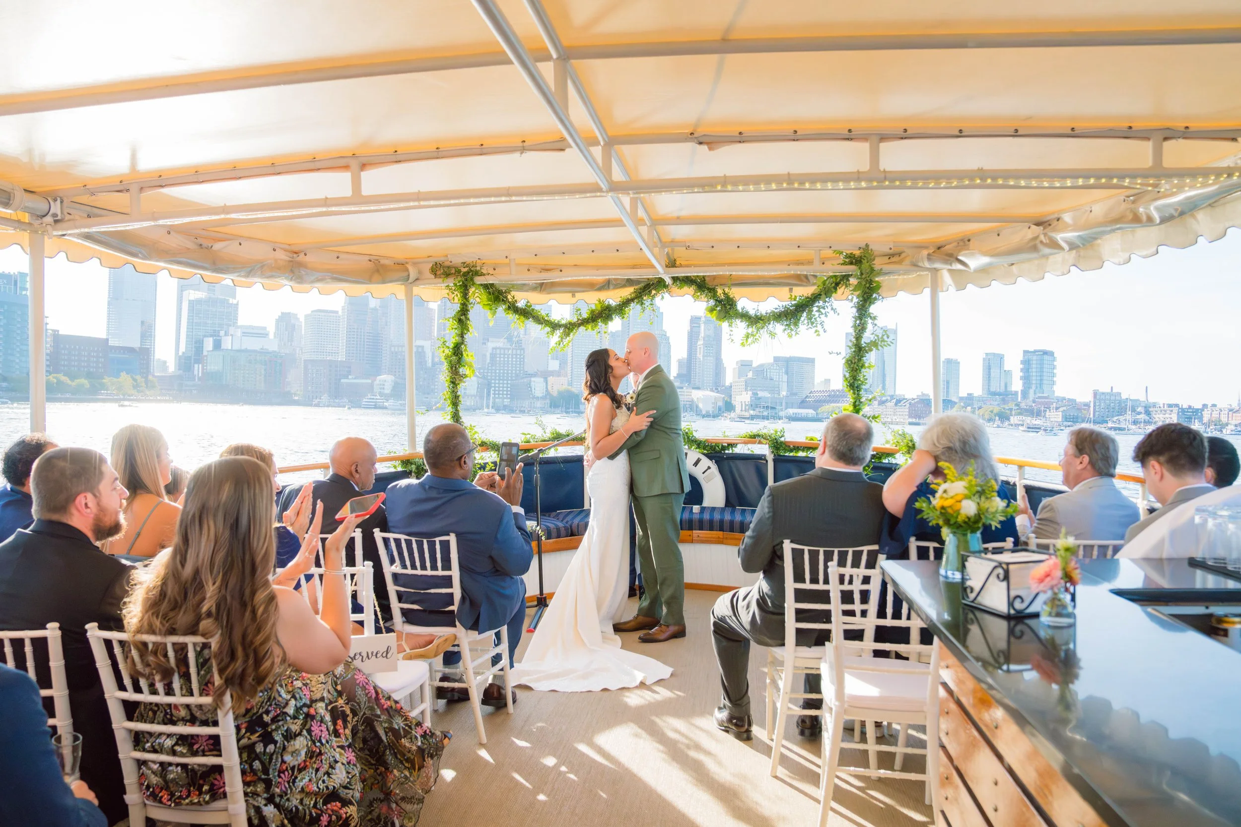 A wedding ceremony on a boat with a city skyline in the background, featuring a bride and groom kissing.