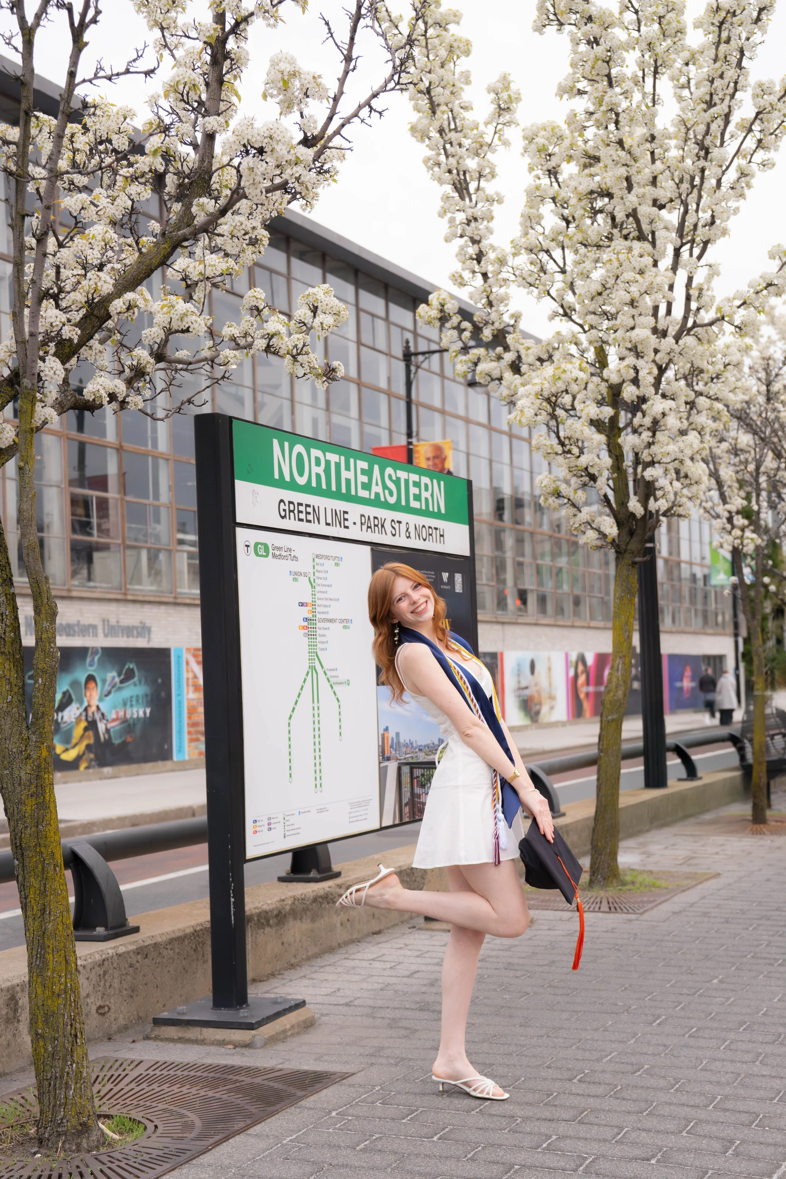 A woman with red hair in a white and navy dress, white high heels, and a necklace, posing playfully on one knee in front of a city bus stop sign at Northeastern University. There are blooming white trees and a modern glass building in the background.