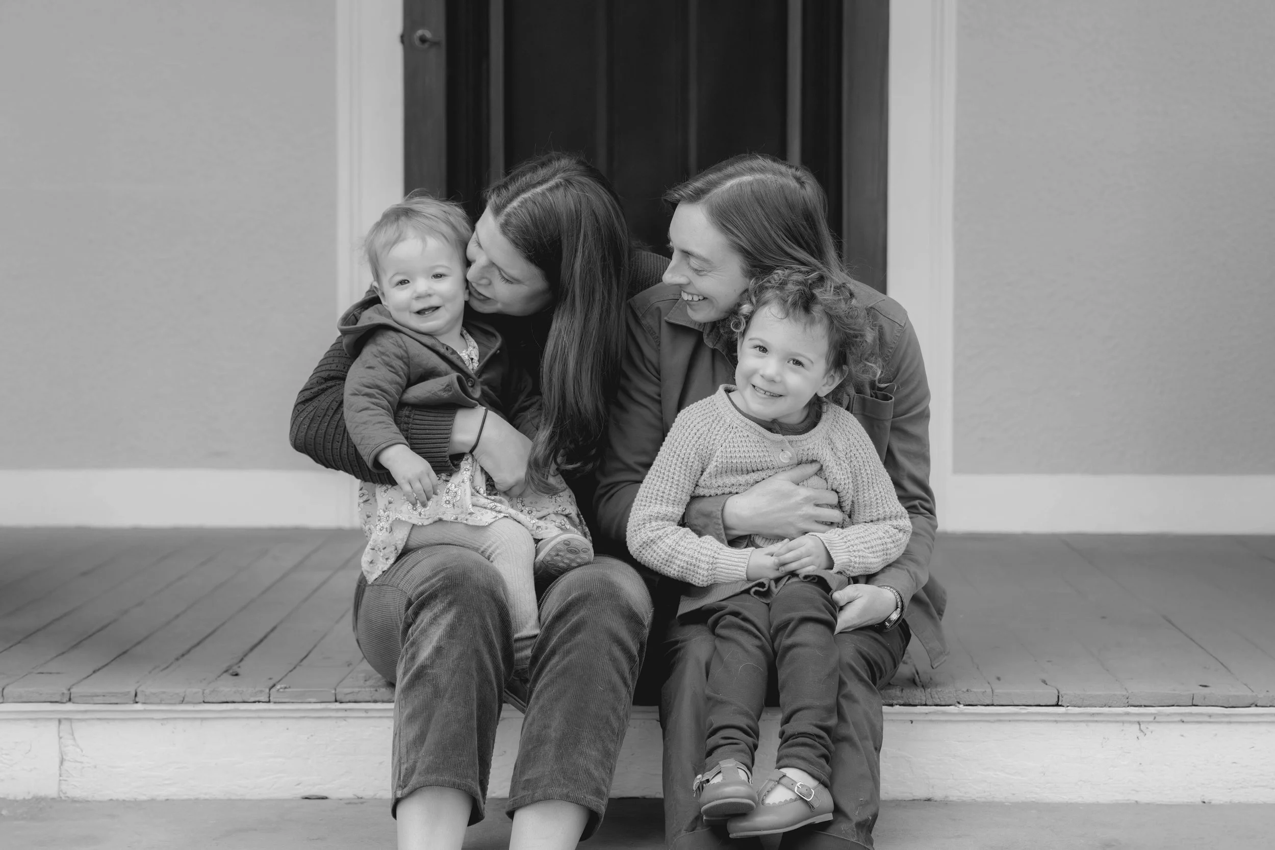 Black and white photo of two women sitting on front steps with two young girls, all smiling and showing affection.