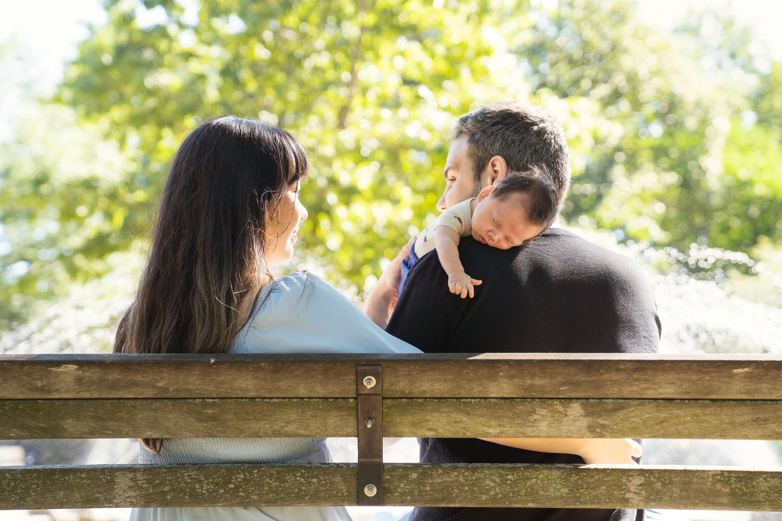 A family of three sitting on a park bench, with a woman, a man, and a baby. The woman and man are looking at each other, and the baby is resting on the man's shoulder. The background is lush and green, indicating a sunny day in a park.