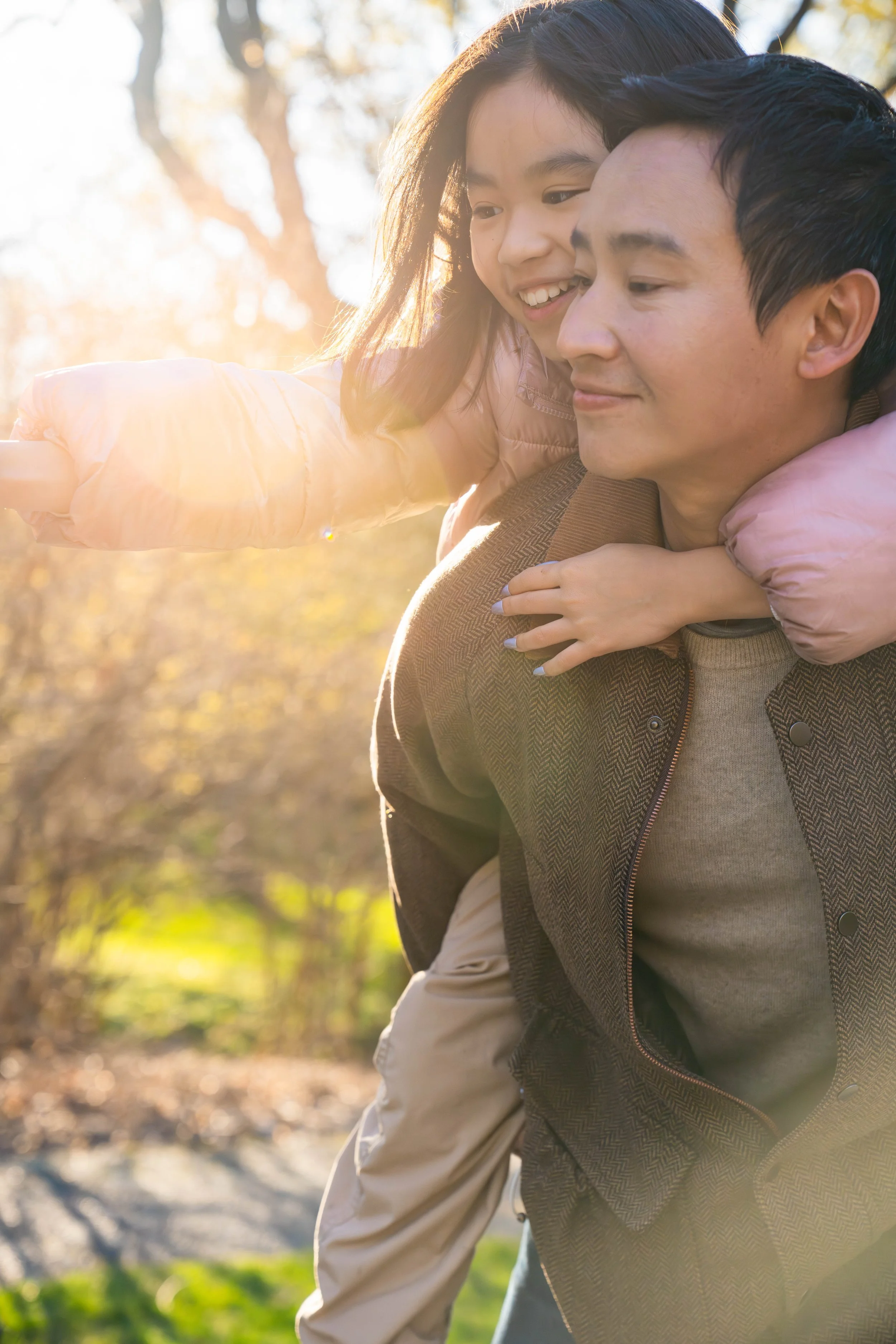 A man giving a young girl a piggyback ride outdoors on a sunny day, with sunlight streaming through the trees.