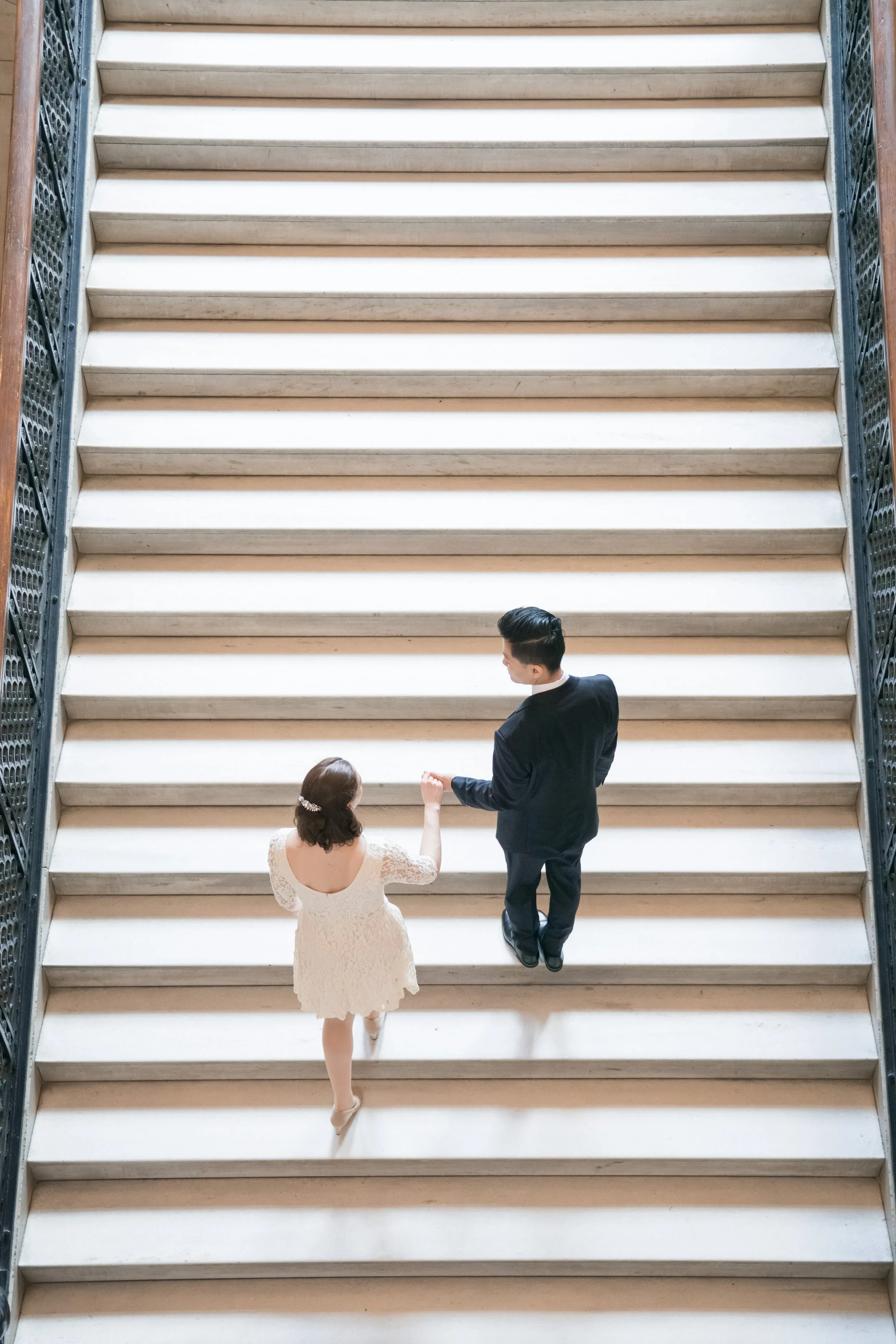 A couple dressed in wedding attire holding hands at the bottom of a large staircase in a grand building.