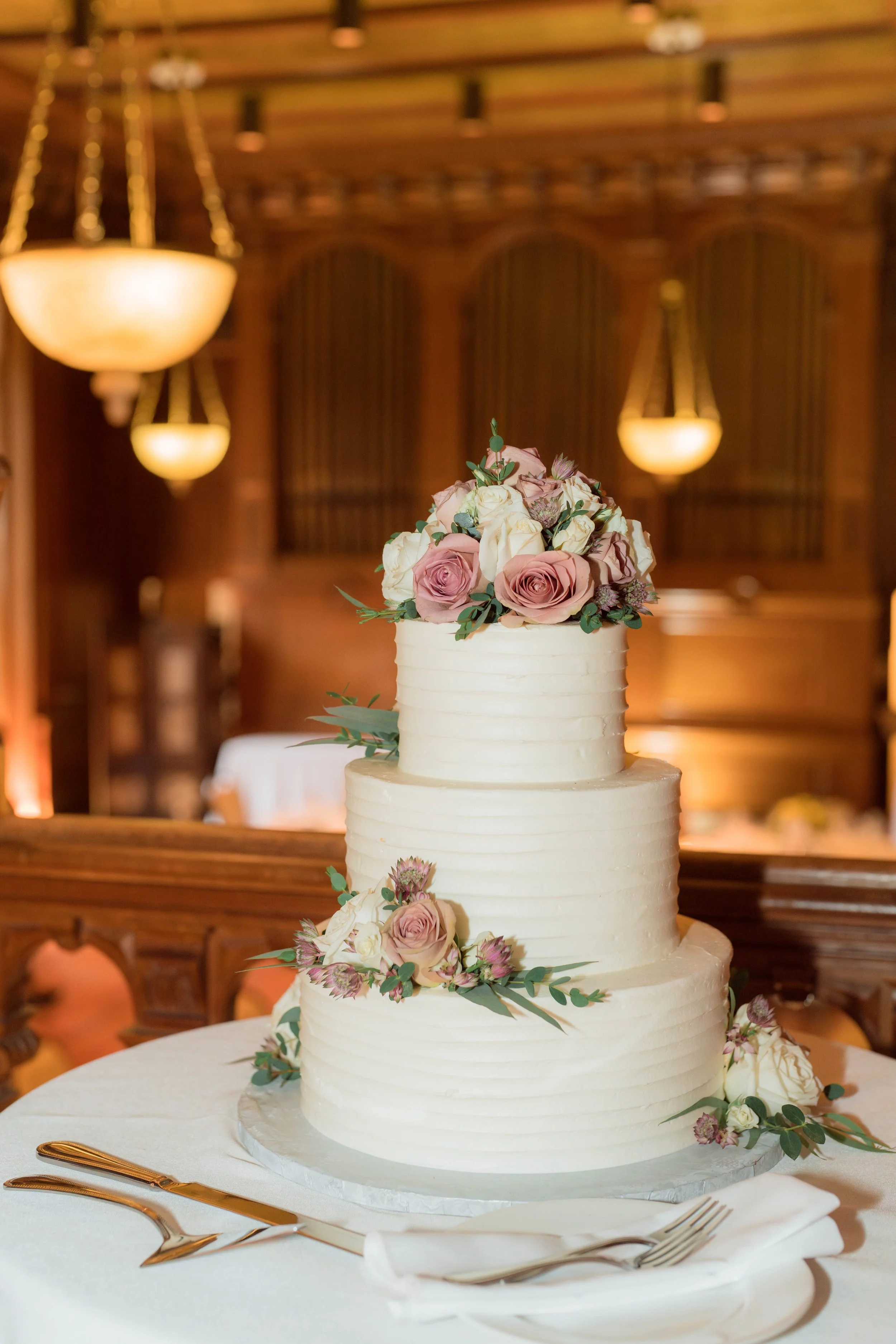 A multi-tiered wedding cake decorated with pink and white roses and greenery, displayed on a table with gold utensils and a white cloth. The background features warm, ambient lighting and wooden paneling.