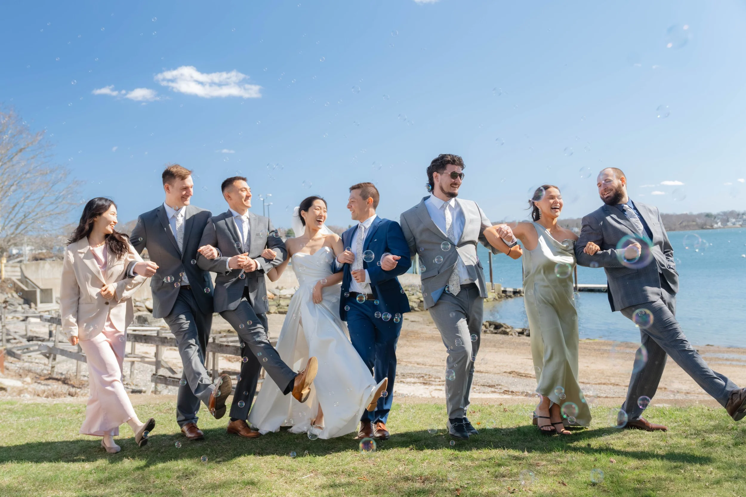 A group of eight people in wedding attire running along a grassy area near water on a sunny day, with bubbles floating in the air.