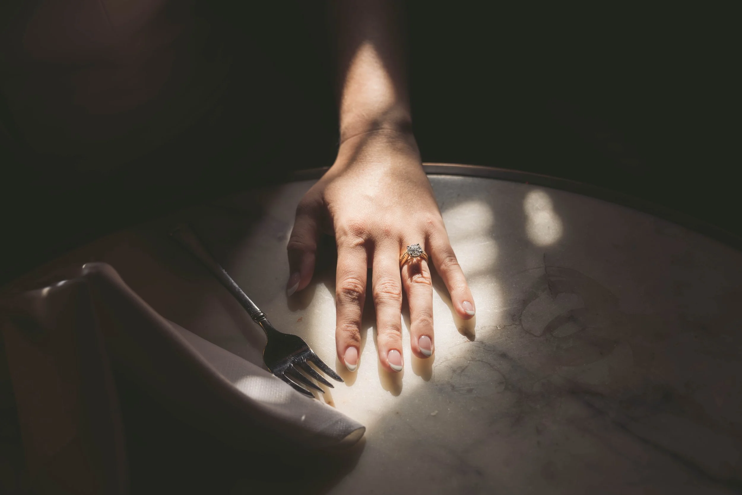 Close-up of a hand with a large gemstone ring, resting on a white surface, with a fork nearby. The background is dark, emphasizing the hand and ring.