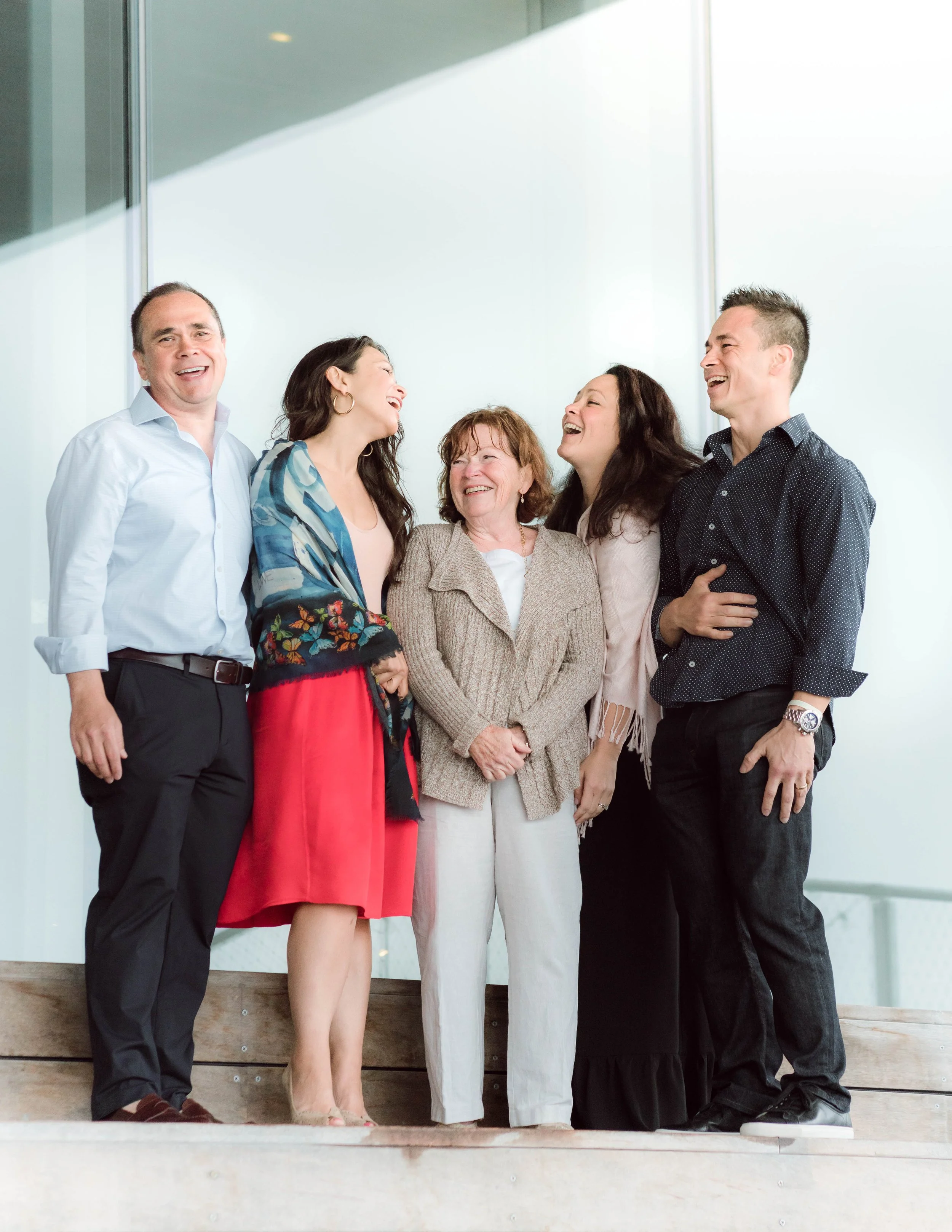 Six people standing together, laughing and smiling in a modern indoor space with a white wall and large window.
