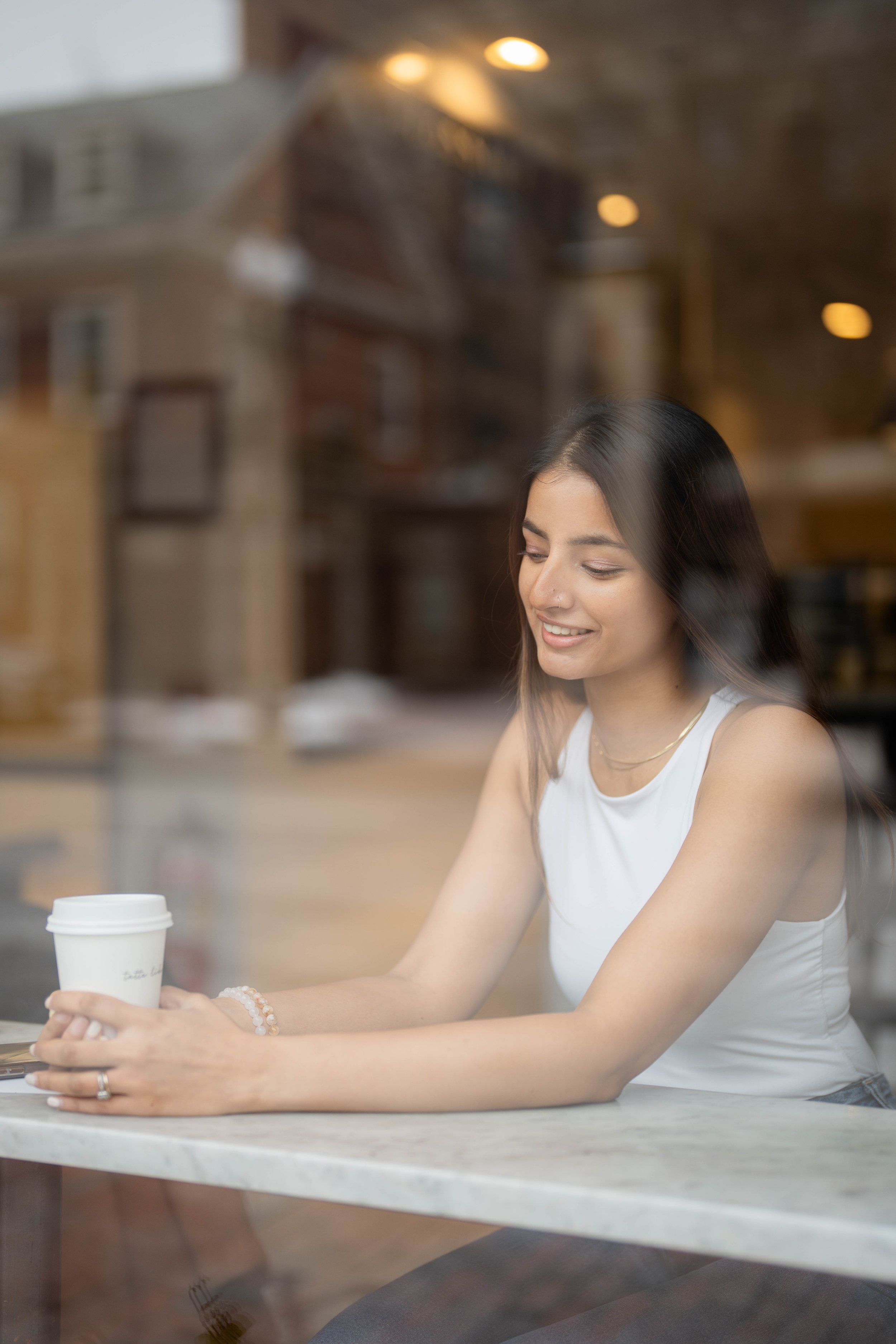 A young woman with long dark hair and a nose piercing sitting at a table in a coffee shop, smiling and holding a white disposable coffee cup with a lid.