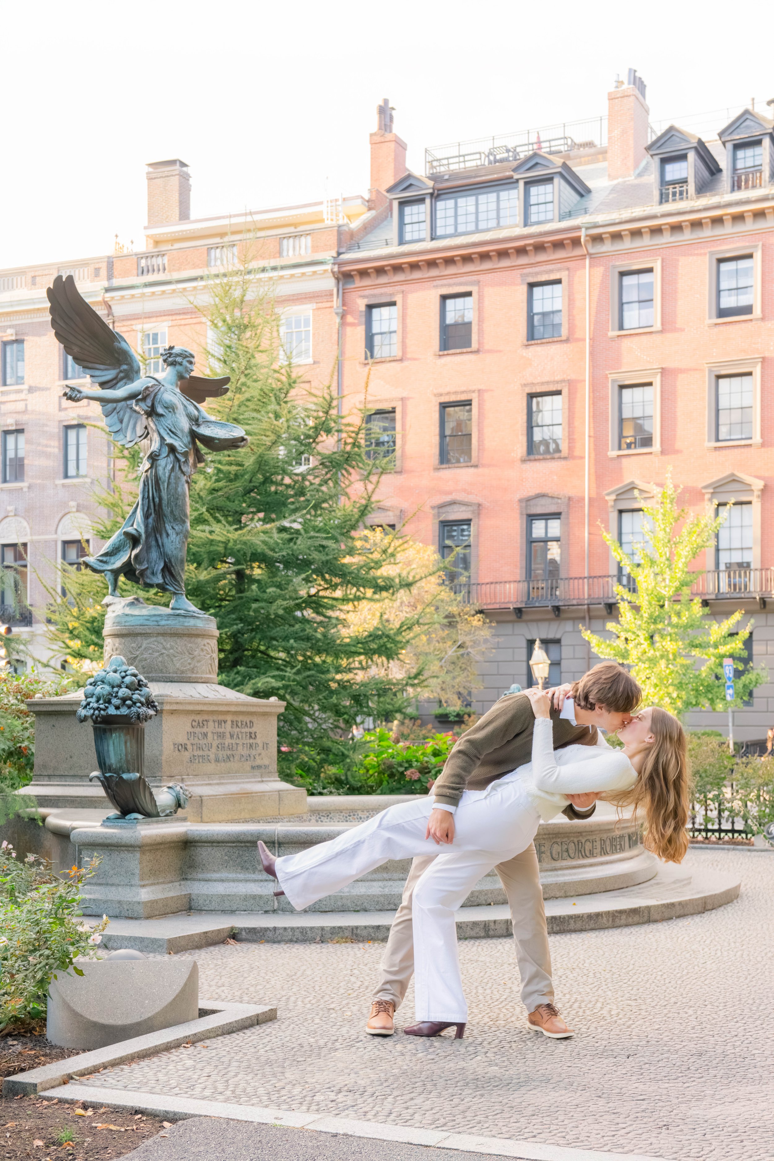 A young man and woman share a kiss in a city park near a fountain with a statue of a woman holding a dish. The man lifts the woman, who is reclined with her arms around his neck. There are brick buildings and trees in the background.