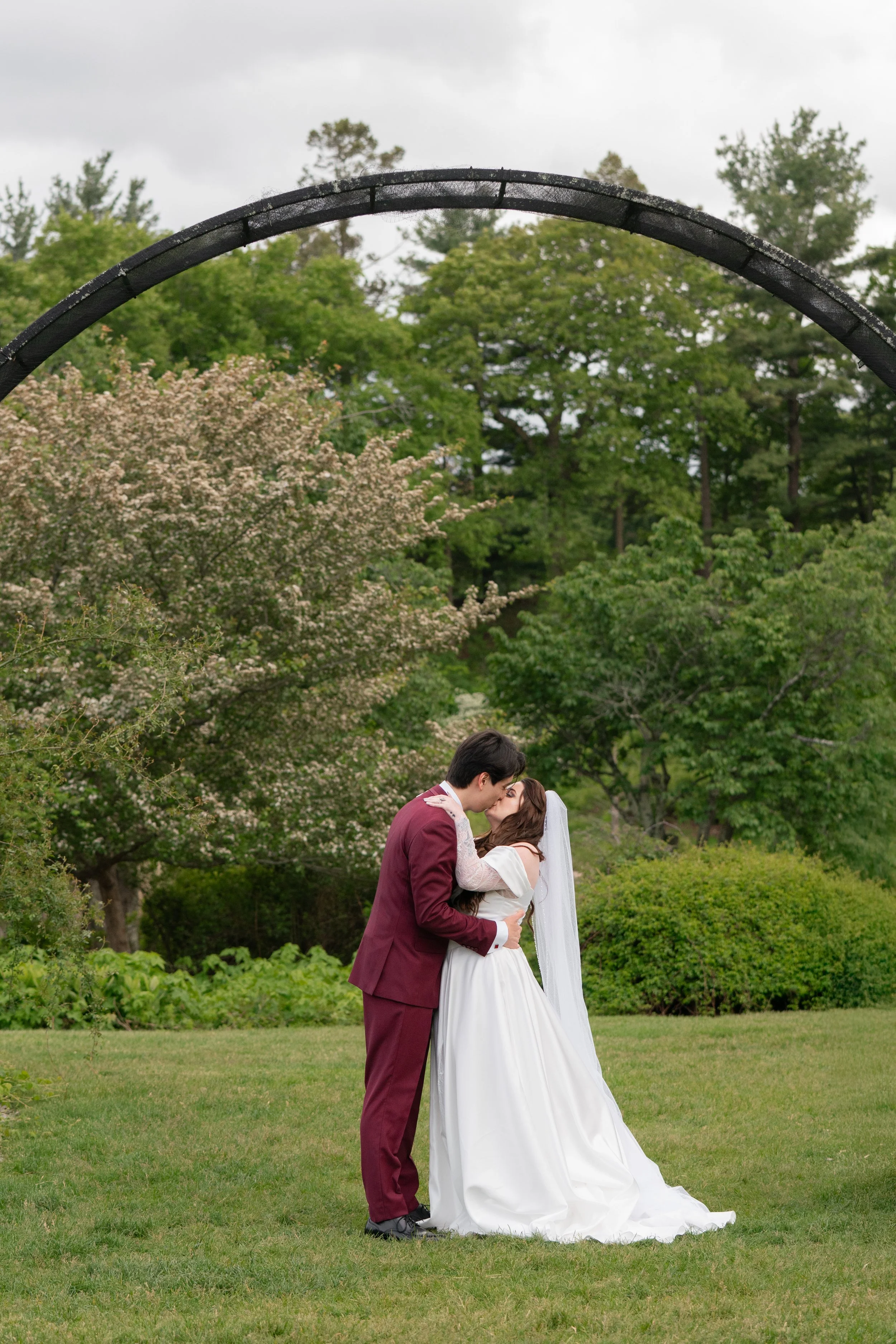 A bride and groom sharing a kiss outdoors in a grassy park with trees and cloudy sky in the background.