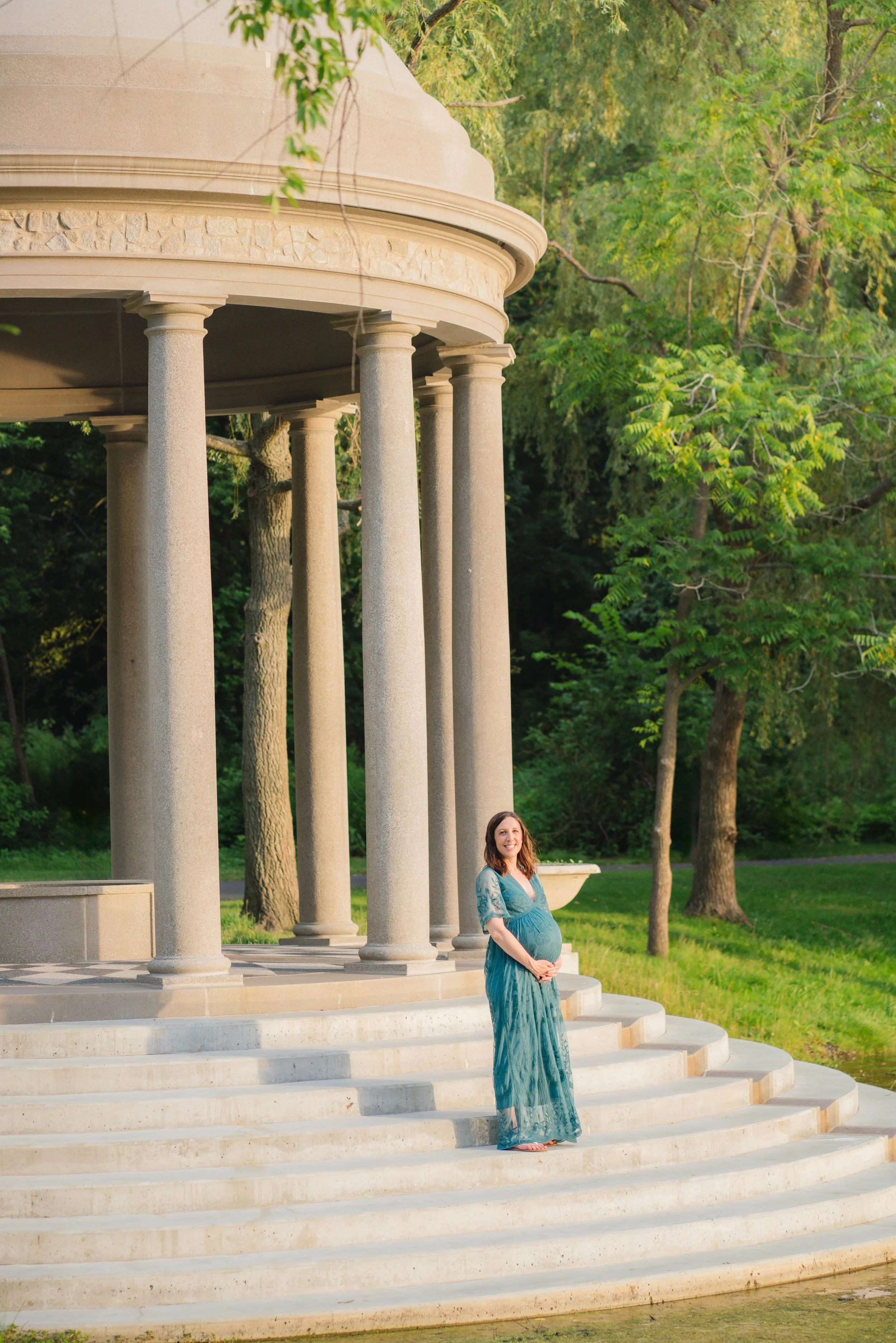 A pregnant woman in a blue dress standing on the steps of a round stone pavilion with columns, surrounded by trees and greenery, smiling and holding her belly.