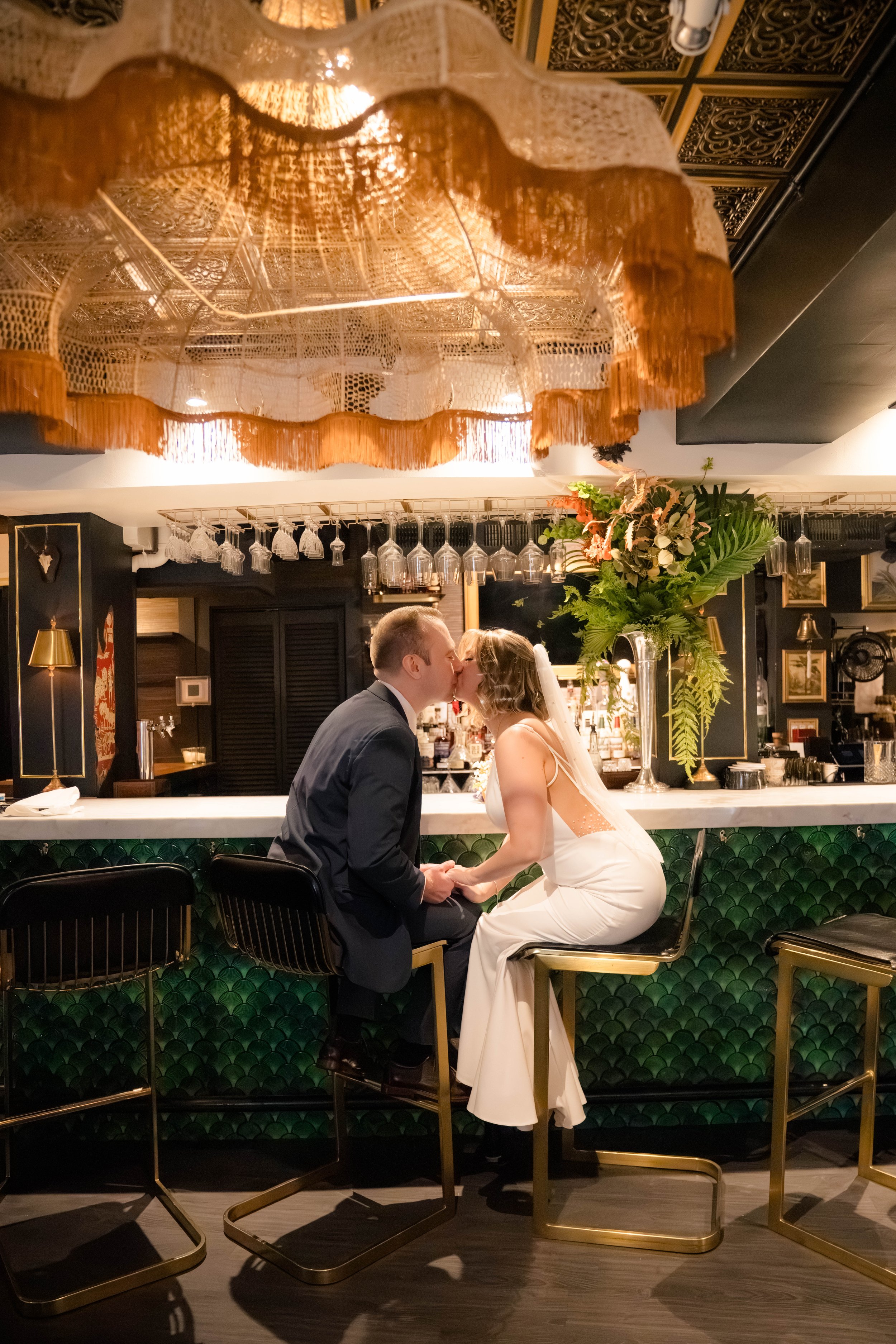 A bride and groom share a kiss while sitting at a bar in an elegant, dimly lit restaurant. The bride is wearing a white wedding dress with a veil, and the groom is dressed in a dark suit. The bar has a green tile front, gold accents, and a large flor