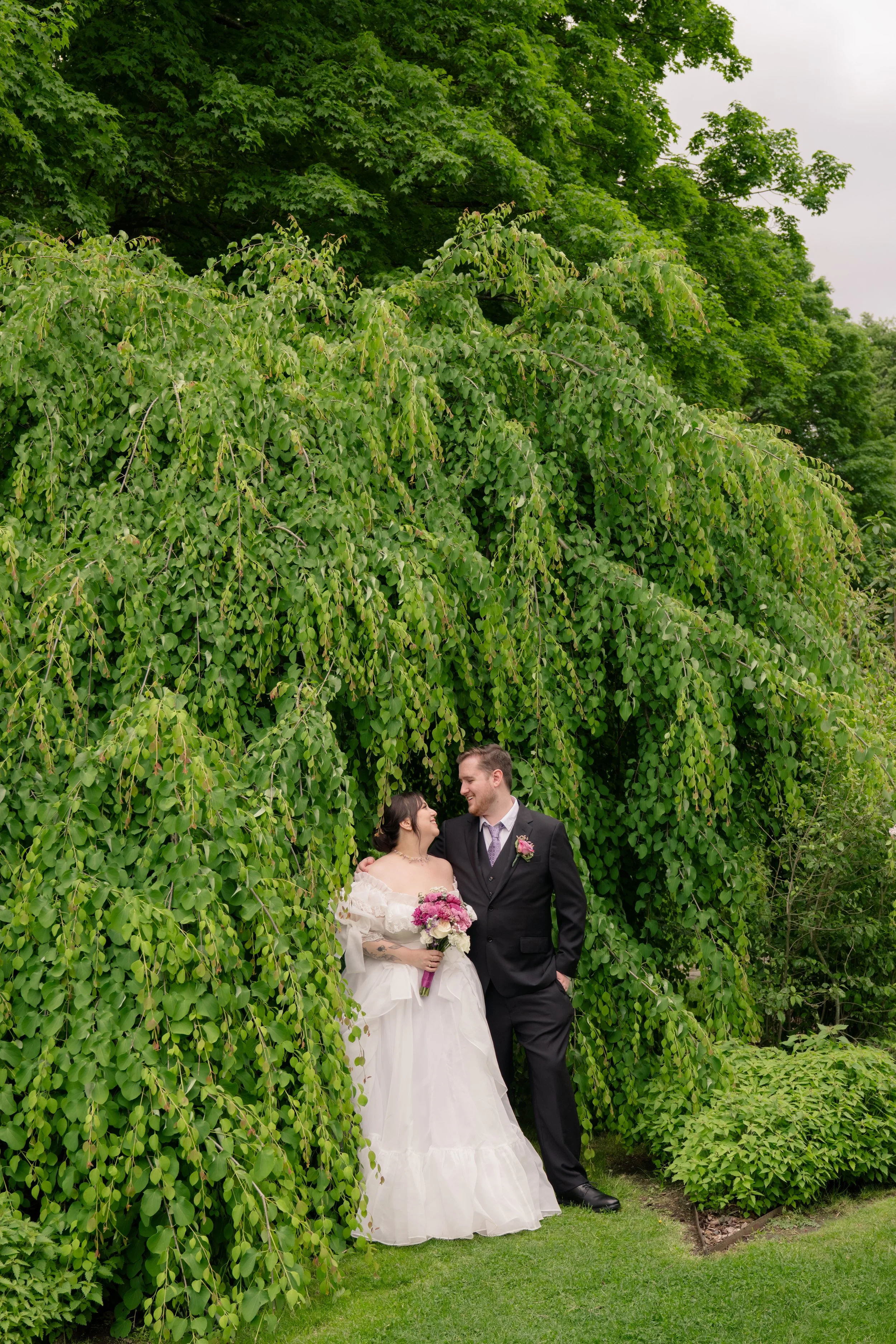 A bride and groom in wedding attire standing beneath a large leafy green arch in a garden, smiling and looking at each other, with the bride holding a bouquet of pink and white flowers.