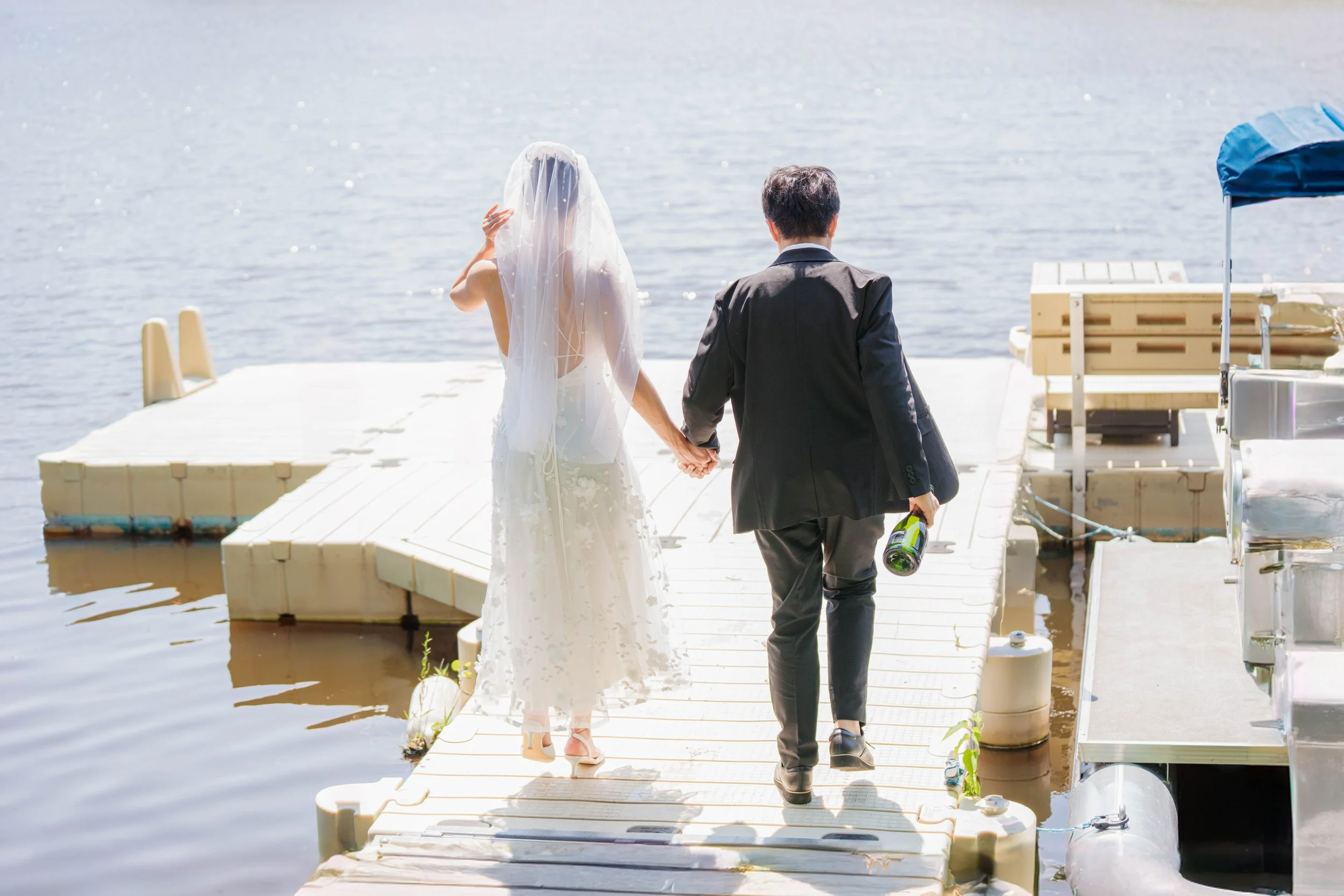 A bride and groom holding hands cross a dock over a lake. The bride is wearing a white wedding dress with a veil, and the groom is dressed in a black suit, holding a bottle of champagne.