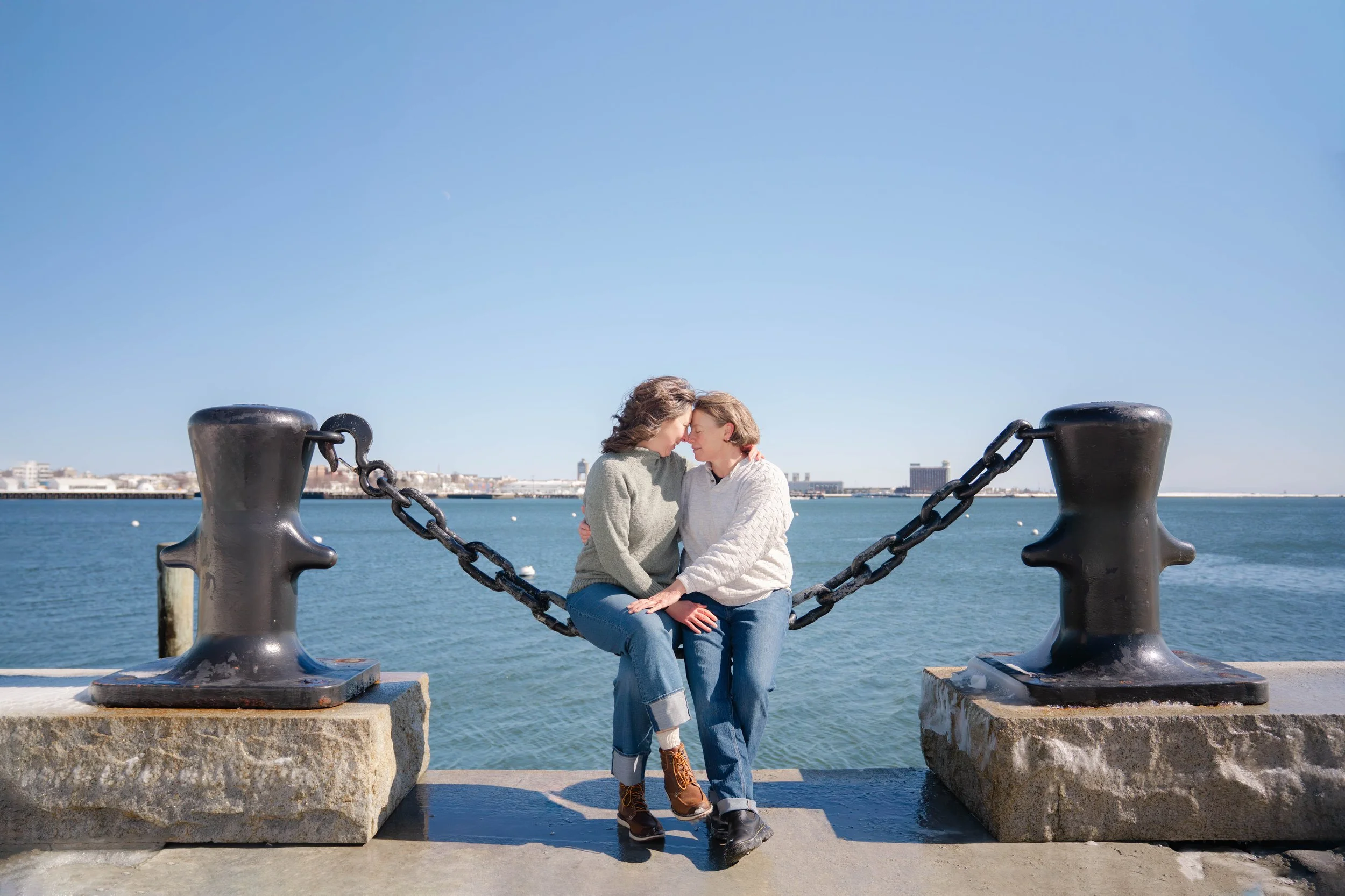 Two women sit closely together on a chain-linked boat docking cleat by the water, with their foreheads touching in an embrace during the day. The background includes a city skyline and a calm body of water under a clear, blue sky.
