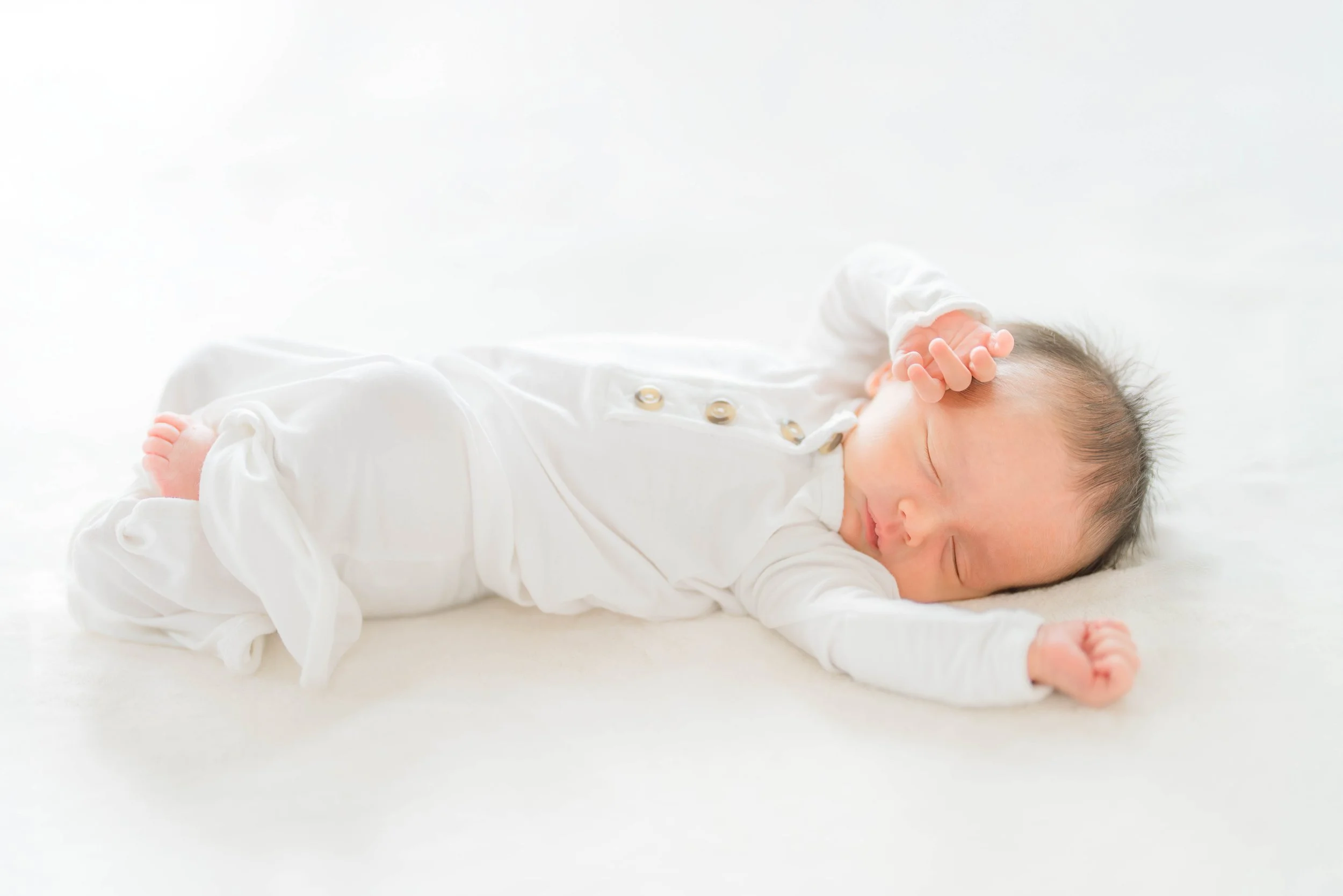 A sleeping baby lying on a white surface, dressed in a white outfit with gold buttons, with a hand resting on the head.