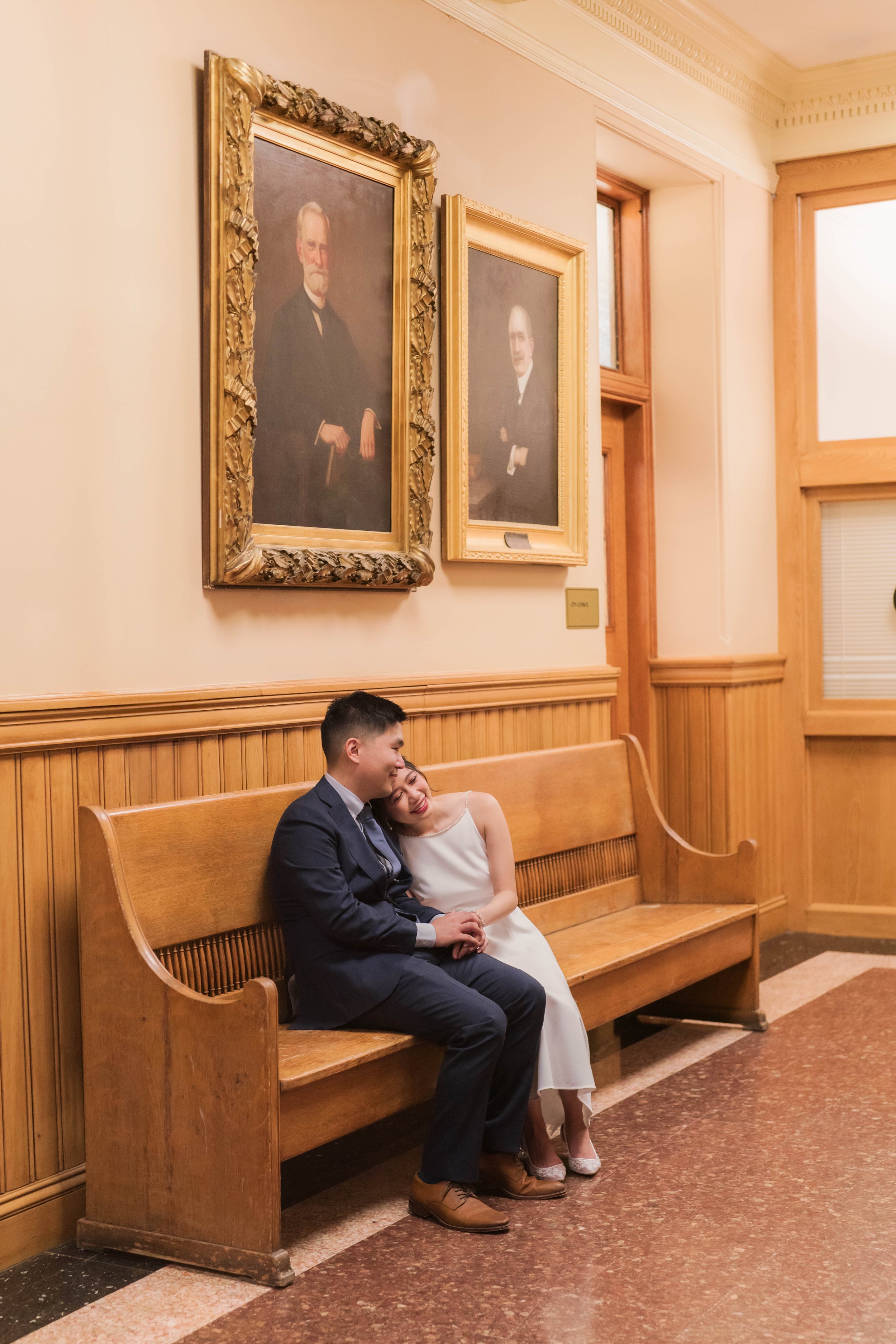 A young couple dressed in formal wedding attire sitting on a wooden bench in a room with wooden-paneled walls, framed portraits hanging on the wall, and large windows with wooden trim.
