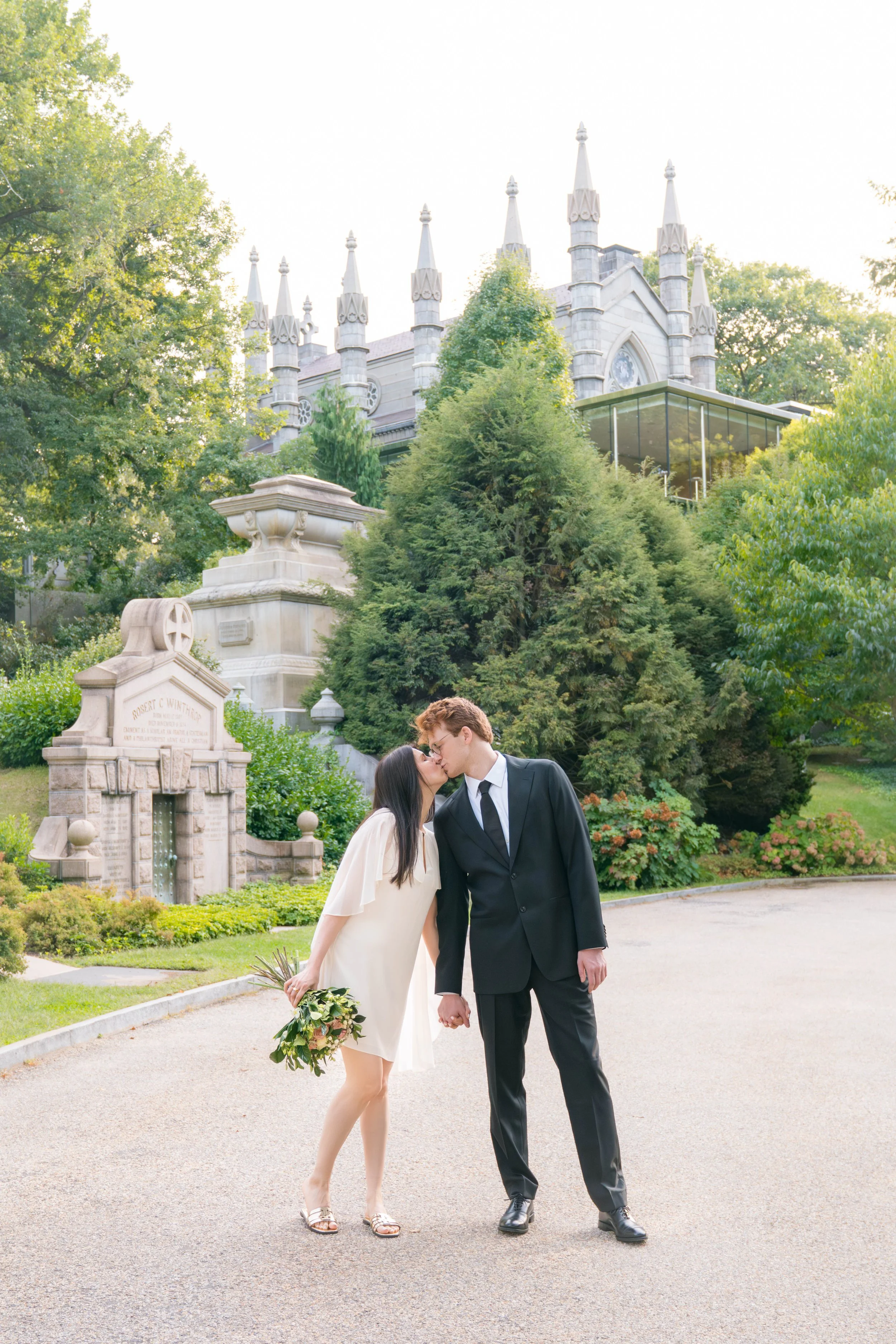A young couple in wedding attire sharing a kiss outdoors in a park with trees and historic buildings in the background.