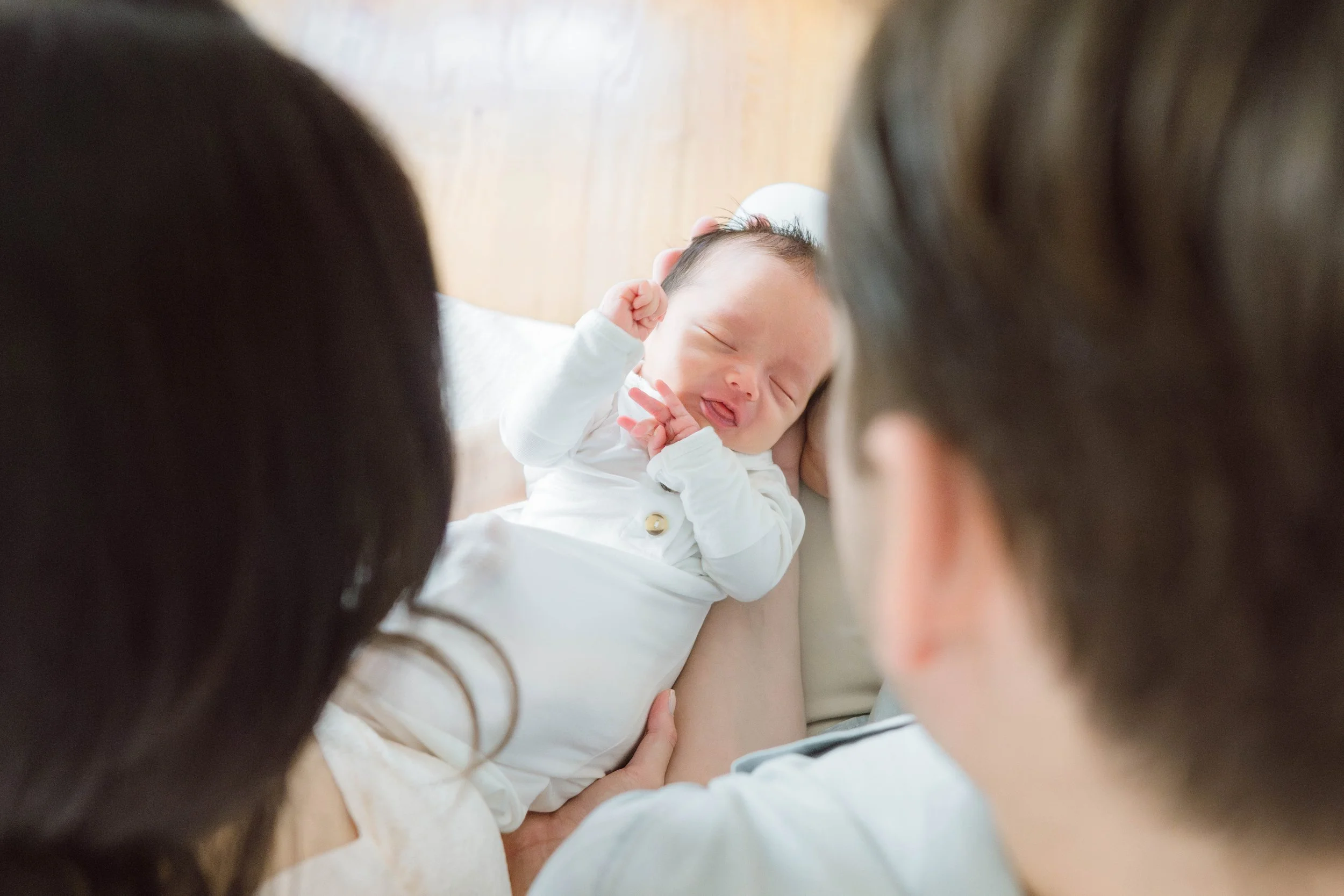 A newborn baby smiling with eyes closed, held by adults who are partly visible around the baby.