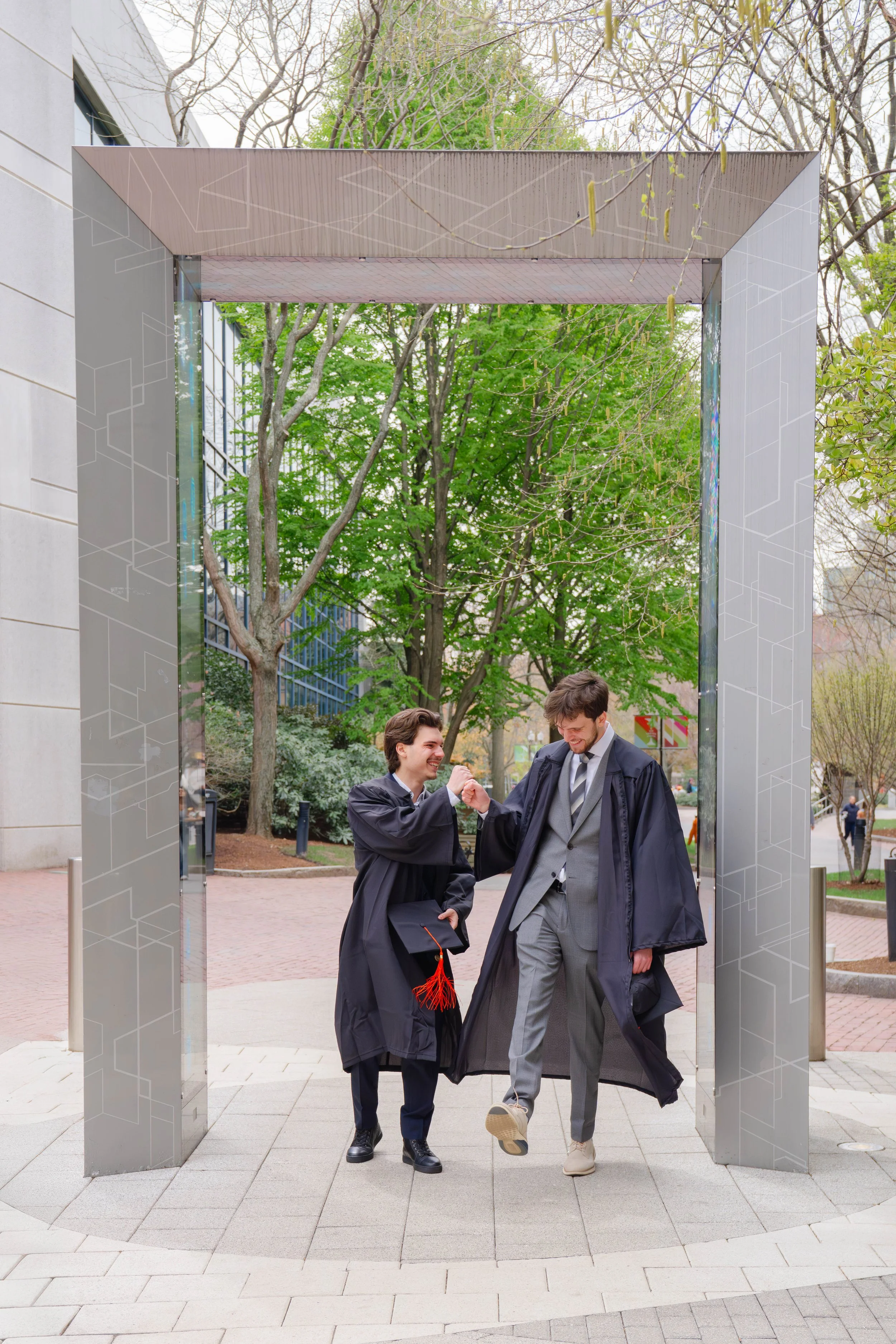 Two young men in graduation gowns are celebrating outside after graduation. One is smiling and holding a graduation cap, and the other is jumping and holding a graduate's stole or necklace. They are under a large metal archway, with trees and a build