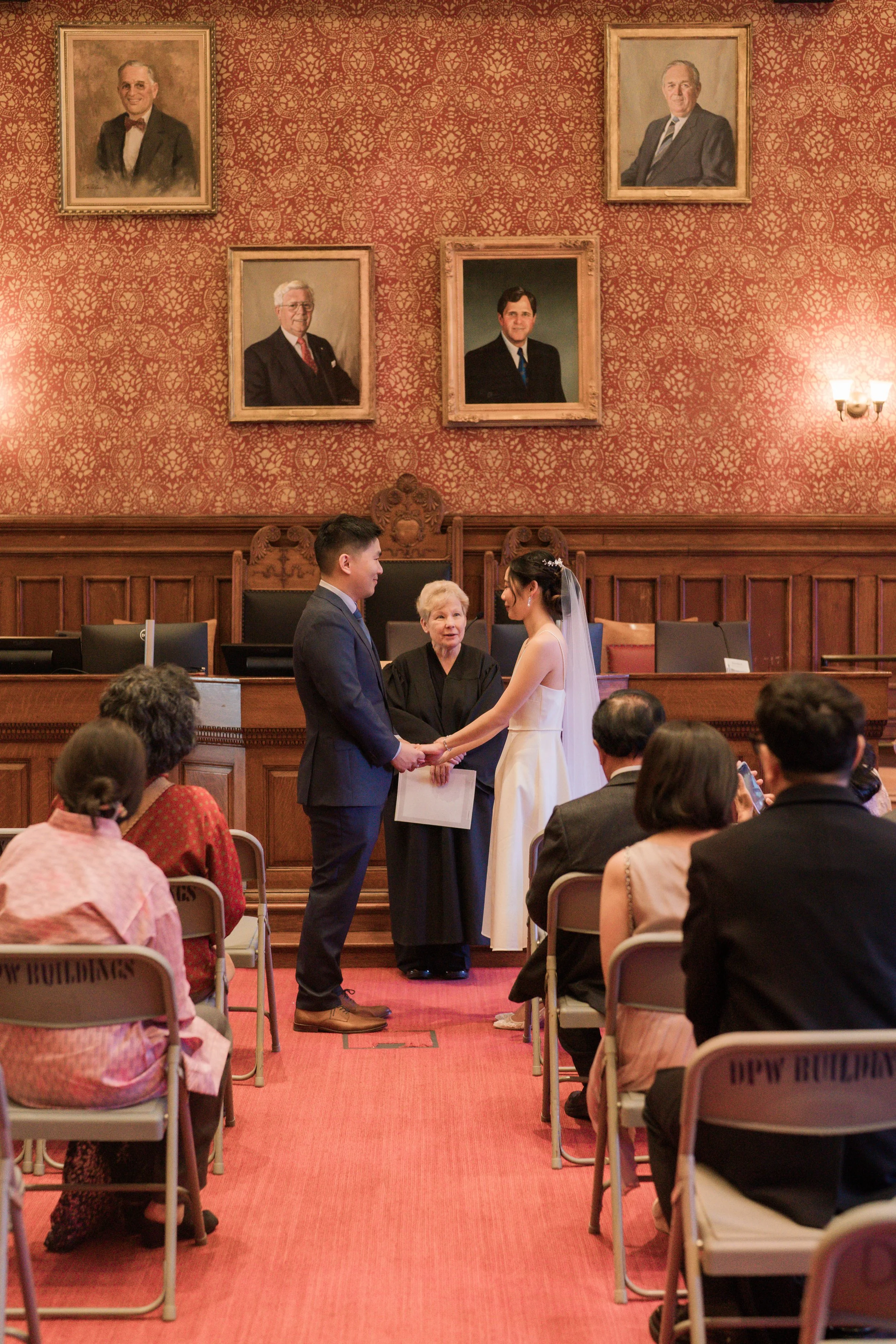 A wedding ceremony taking place in a formal room with ornate red wallpaper and framed portraits on the wall. A bride and groom are holding hands in front of a female officiant, while guests are seated and observing.