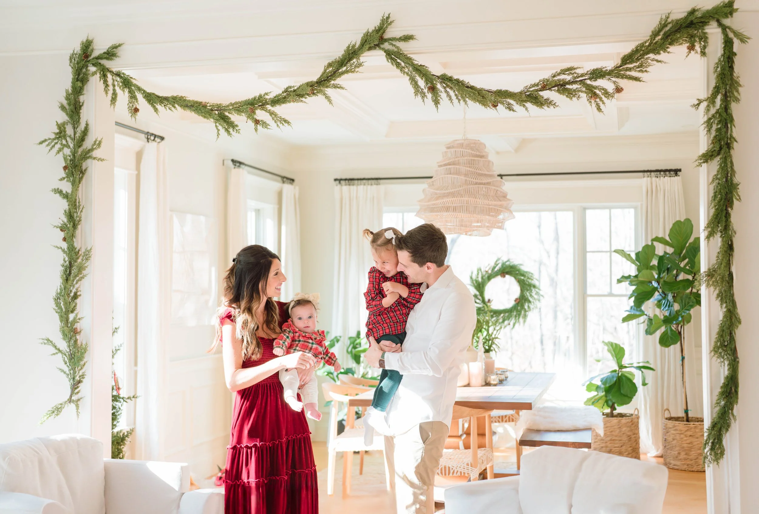 A family of four celebrating Christmas in a bright living room decorated with green garlands hanging from the ceiling and large windows with white curtains. The mother, father, and two children are smiling and interacting with each other.