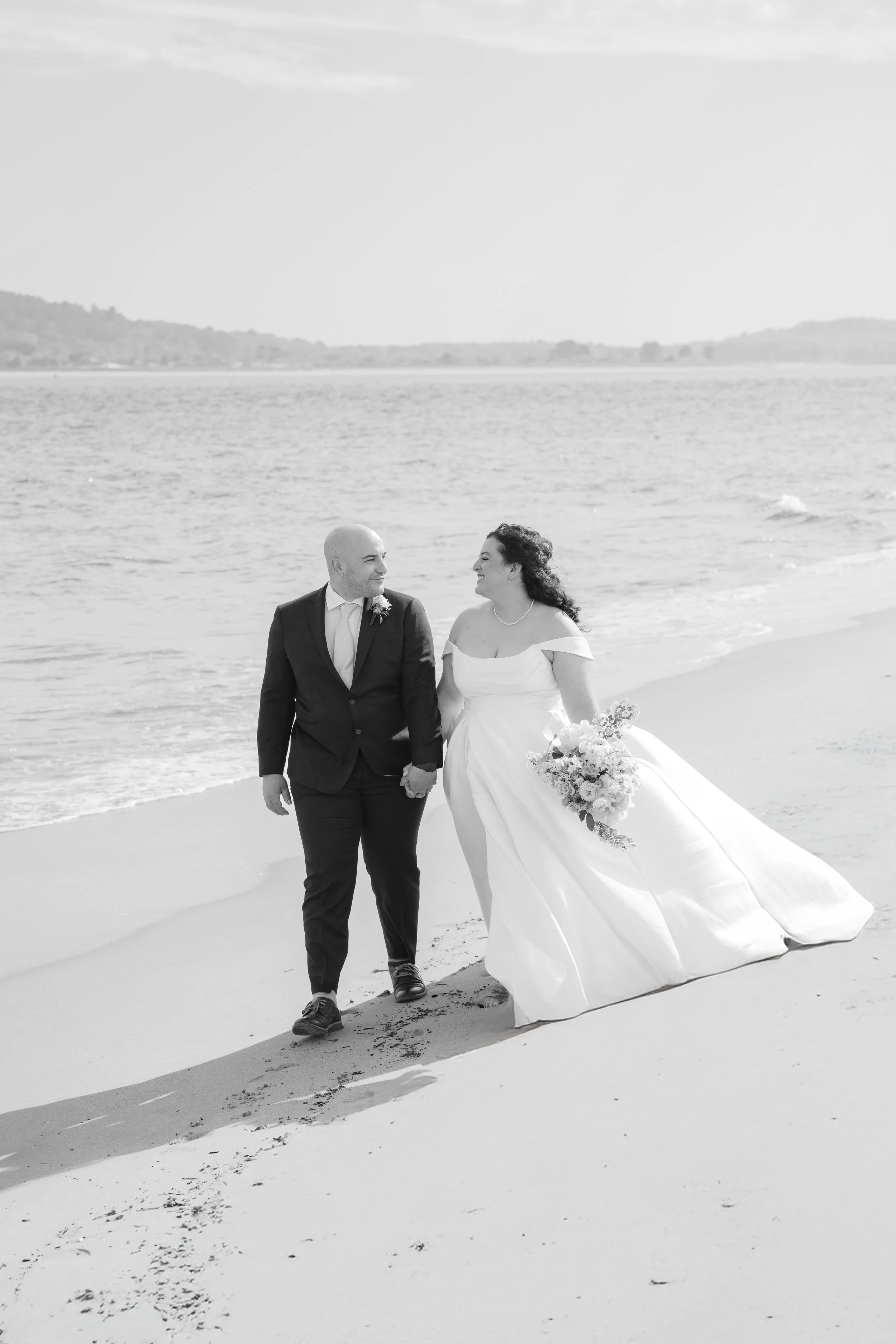 A bride and groom walking hand in hand along the beach, holding hands and smiling at each other. The bride is wearing a white wedding gown and the groom is in a suit. The bride is holding a bouquet of flowers. The ocean and distant hills are visible 