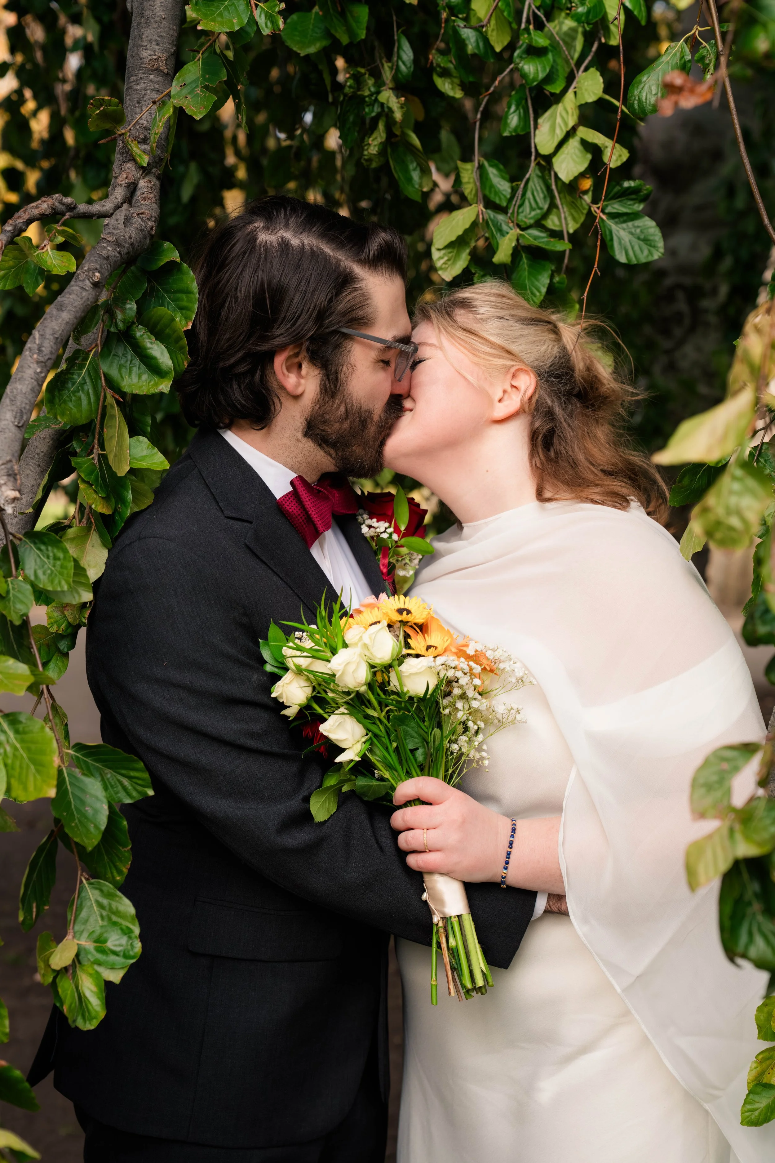 A couple in wedding attire sharing a kiss outdoors, surrounded by green foliage, with the woman holding a bouquet of flowers.