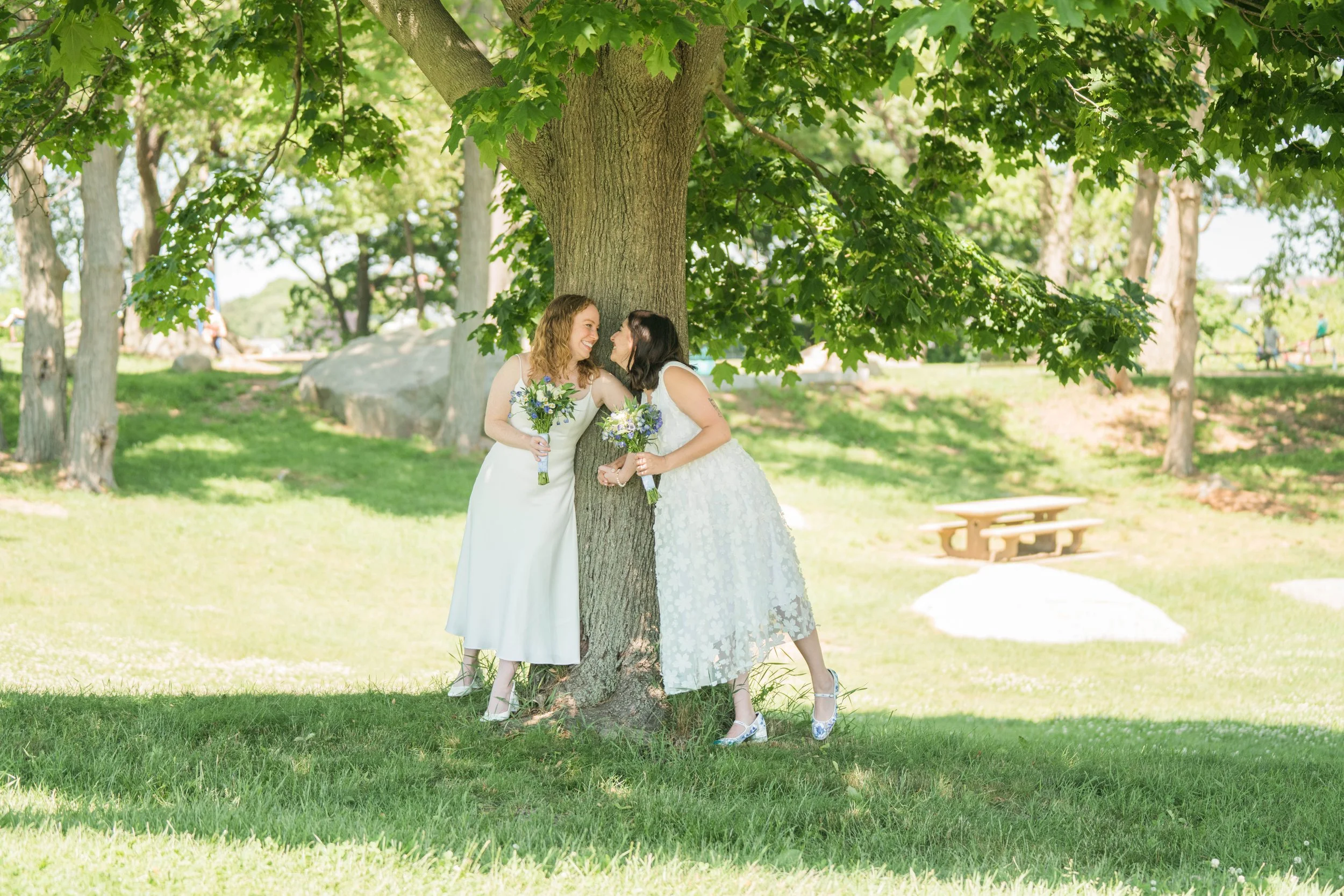 Two brides in white dresses smiling and leaning against a tree, holding bouquets, in a park with green grass and trees.