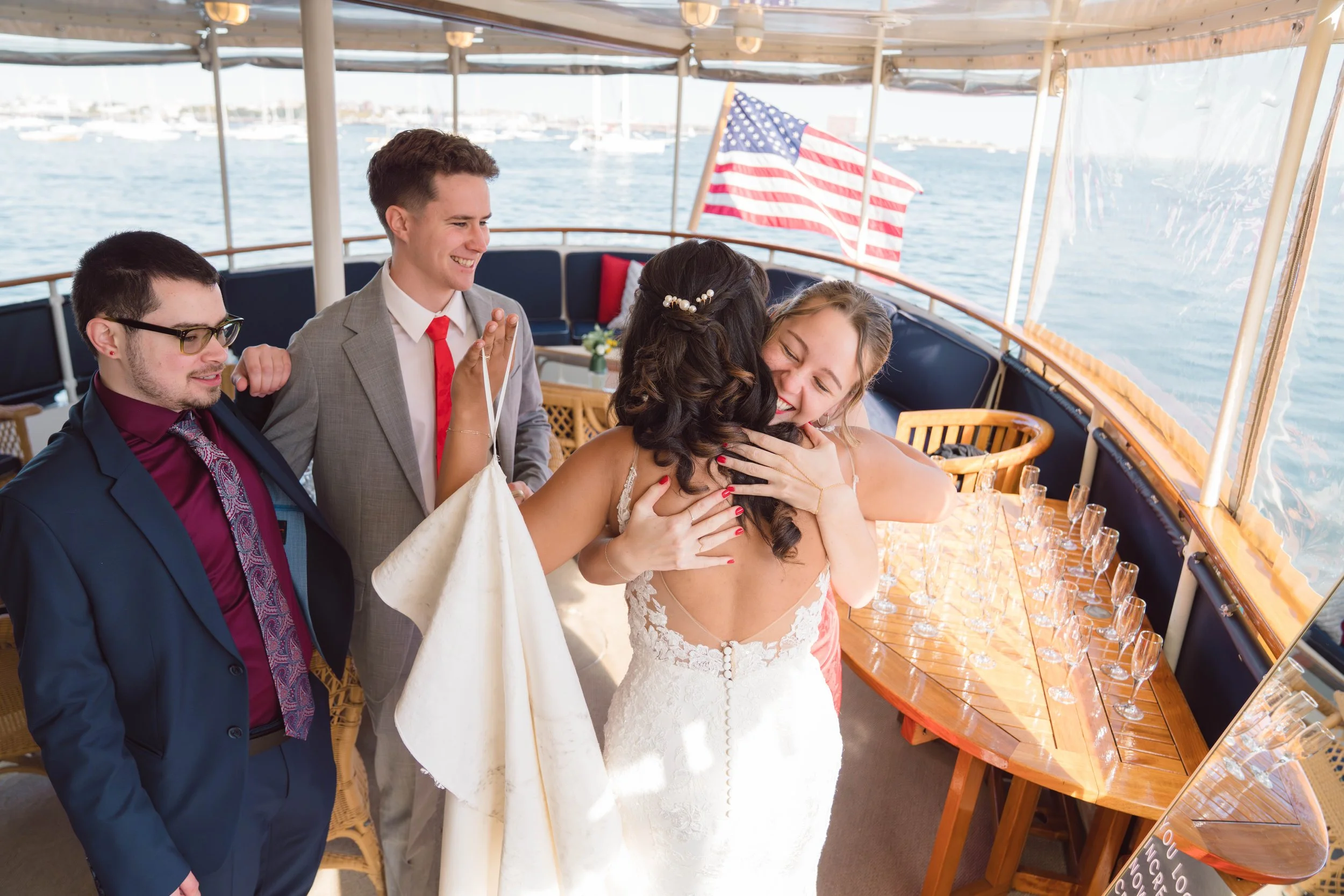 A bride and groom hugging on a boat during their wedding celebration with friends, American flags, and a waterfront view.