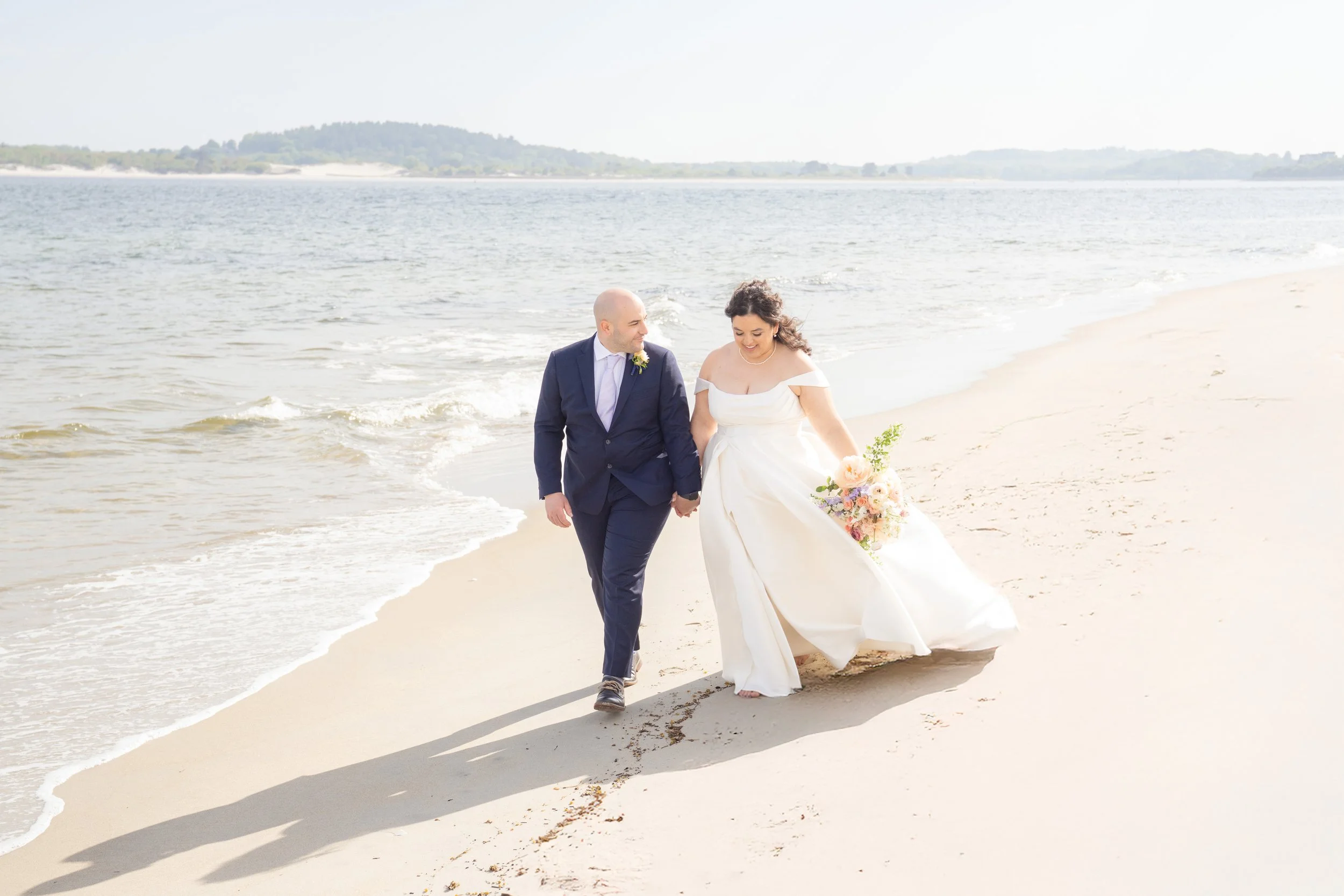 A newlywed couple walking hand in hand along the beach shoreline during their wedding day, with the ocean and distant land in the background.