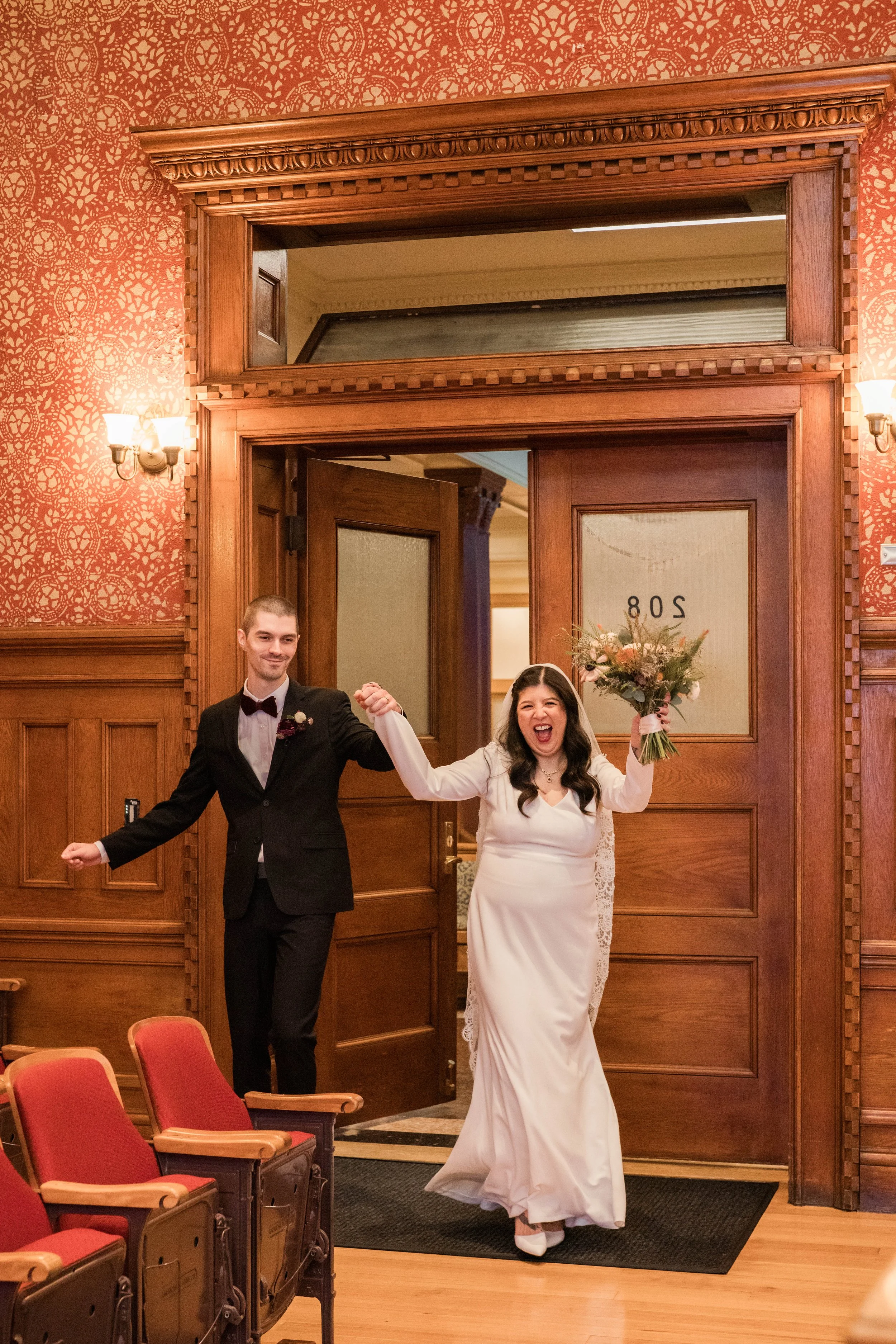 A bride and groom holding hands and walking through a doorway, smiling. The groom is wearing a black tuxedo and the bride is in a white wedding dress holding a bouquet. The room has wood paneling and ornate decor.