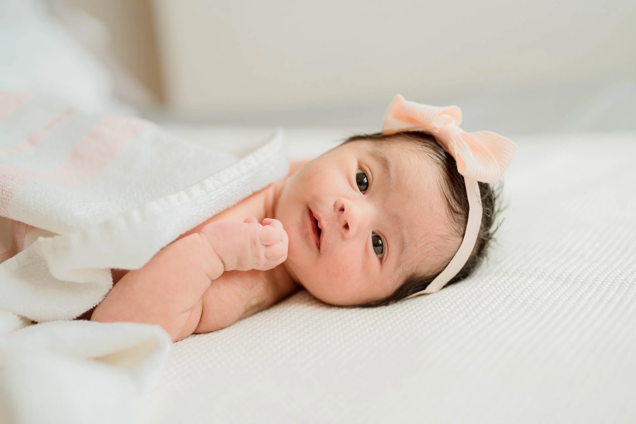 Close-up of a smiling baby girl with dark hair, lying on a white blanket, wearing a peach-colored bow headband and a white outfit.