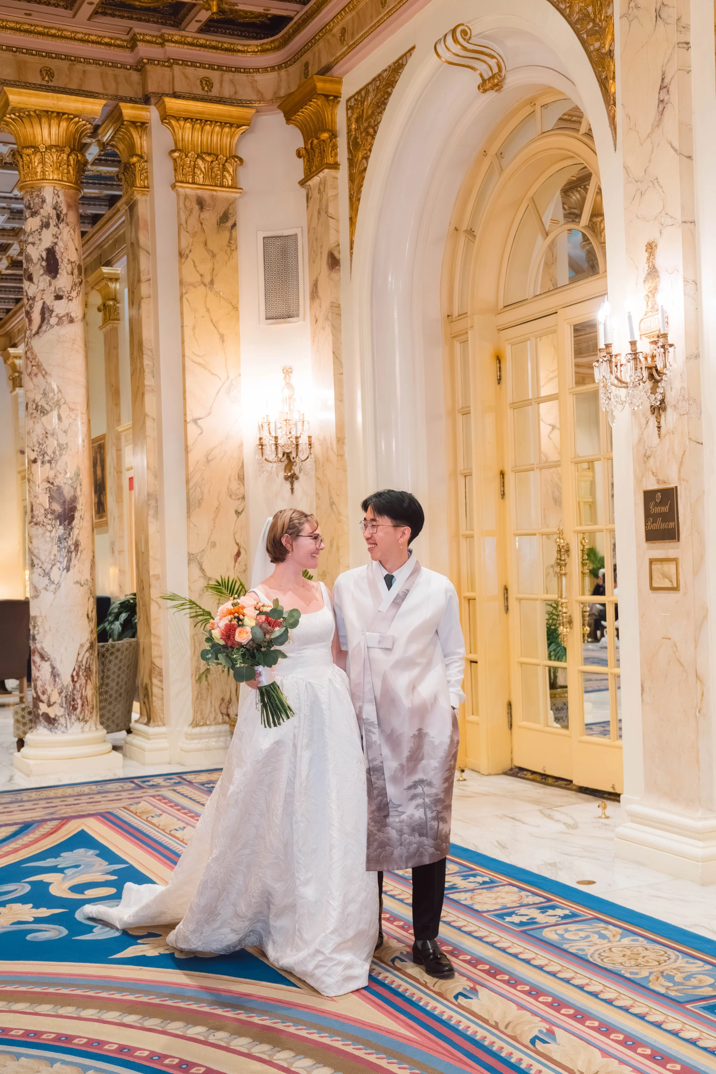 A newlywed couple stands in a grand, ornate ballroom with gold-trimmed marble columns and high arched windows. The bride holds a bouquet of pink and orange flowers, and the groom is dressed in a traditional Korean wedding hanbok. They are smiling at 