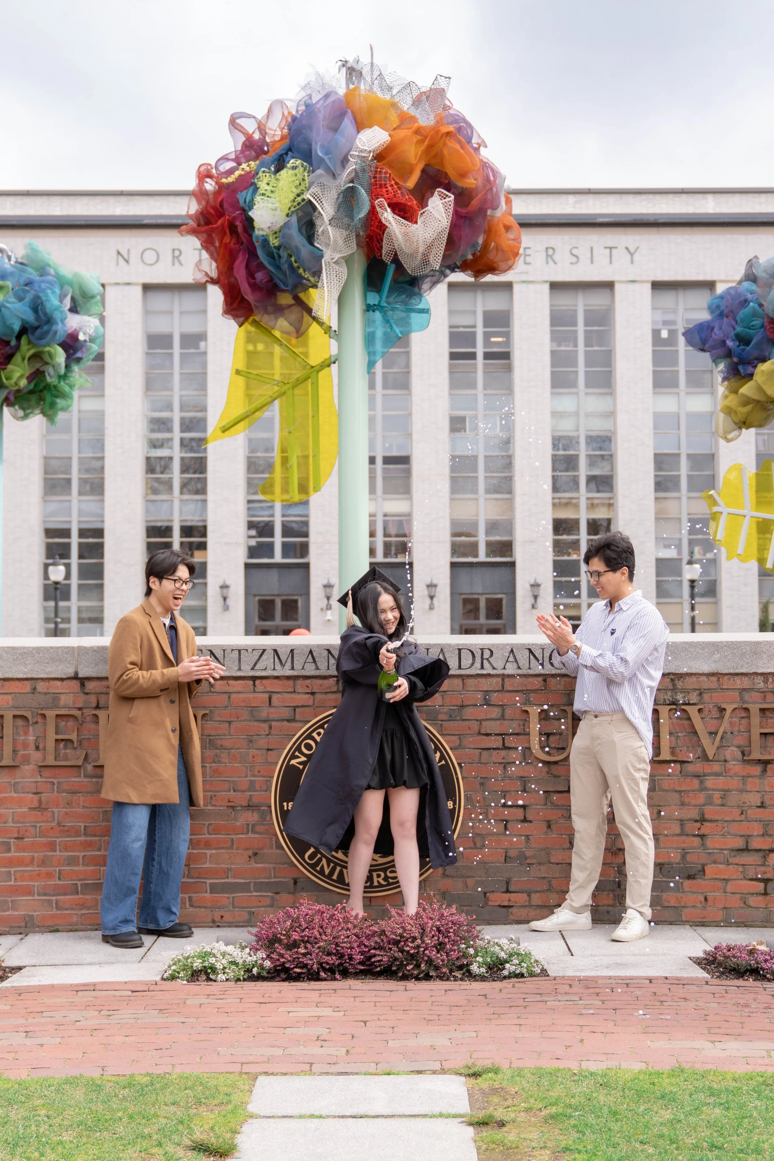 A group of three young people celebrating graduation outdoors in front of a university building. The graduate, in a black gown and cap, stands at the center holding a champagne bottle. Two friends on either side, clapping and smiling. Colorful decorations, including large fabric flowers and leaves, are on the lampposts above them. Flowers are planted in front of a brick wall with a university emblem.