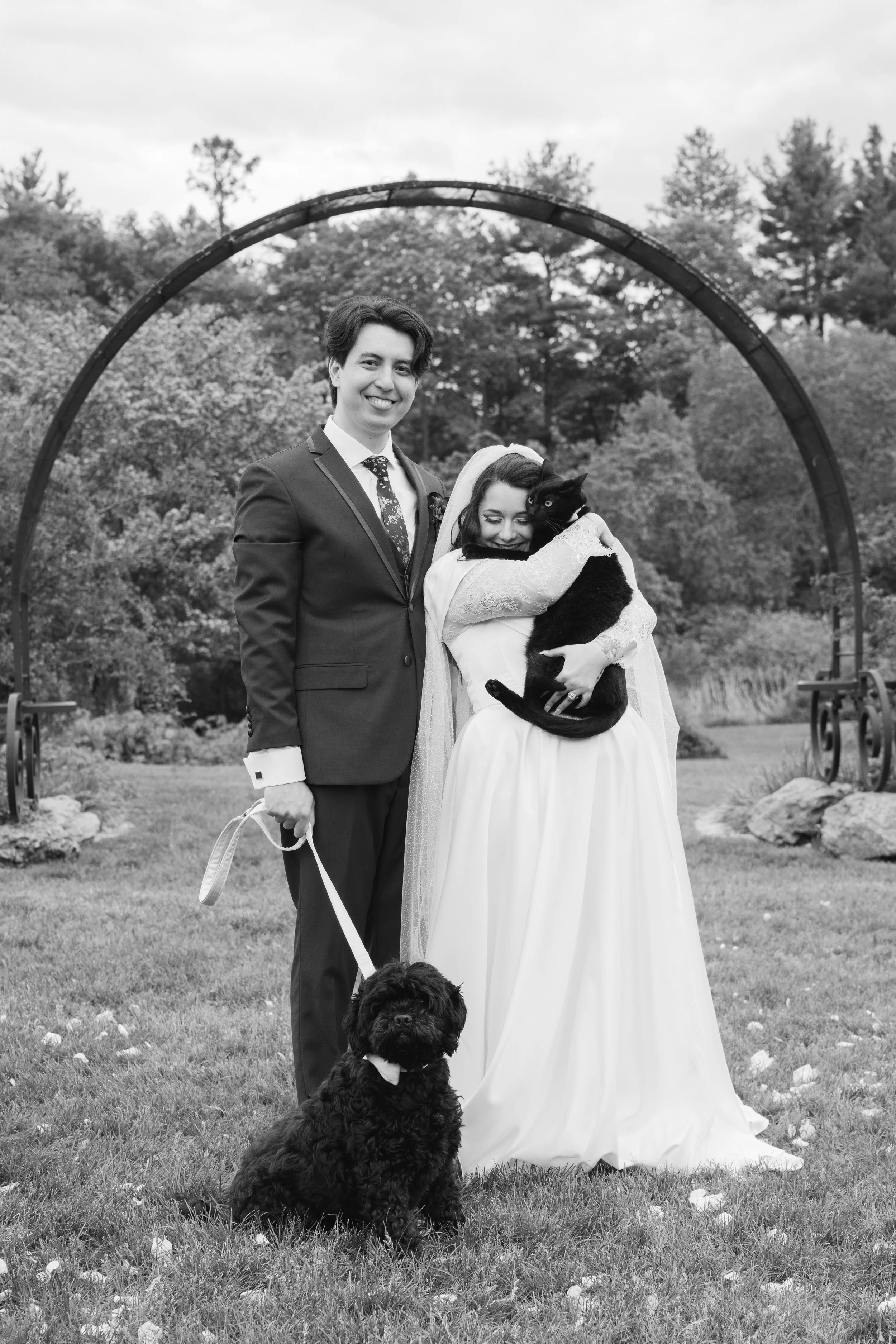 Black and white photograph of a happy bride and groom outdoors, with two dogs. The groom is in a suit and the bride is in a wedding dress, hugging a black dog. A larger black dog sits on the grass in the foreground. Trees and a metal arch are in the 