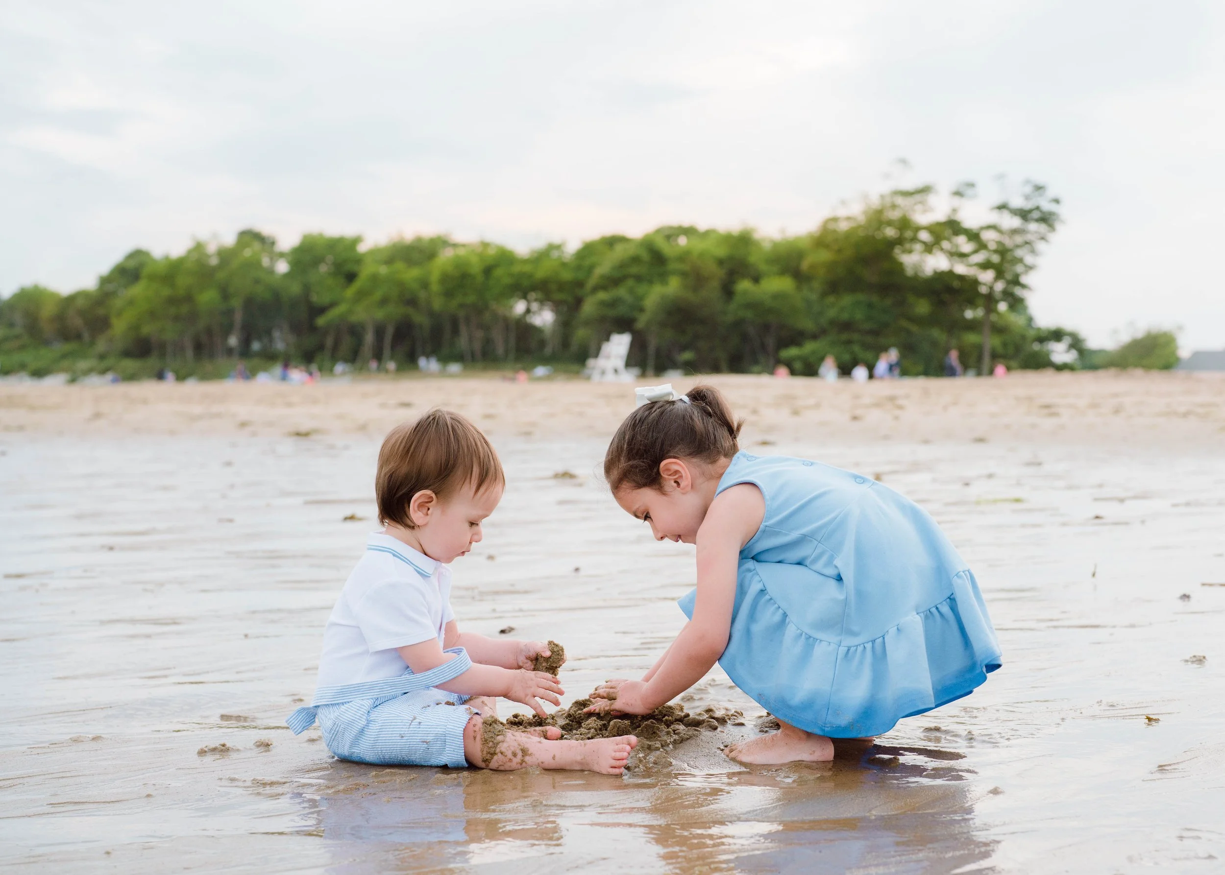 Two young children playing in the sand on a beach, with a green forest in the background, under a cloudy sky.