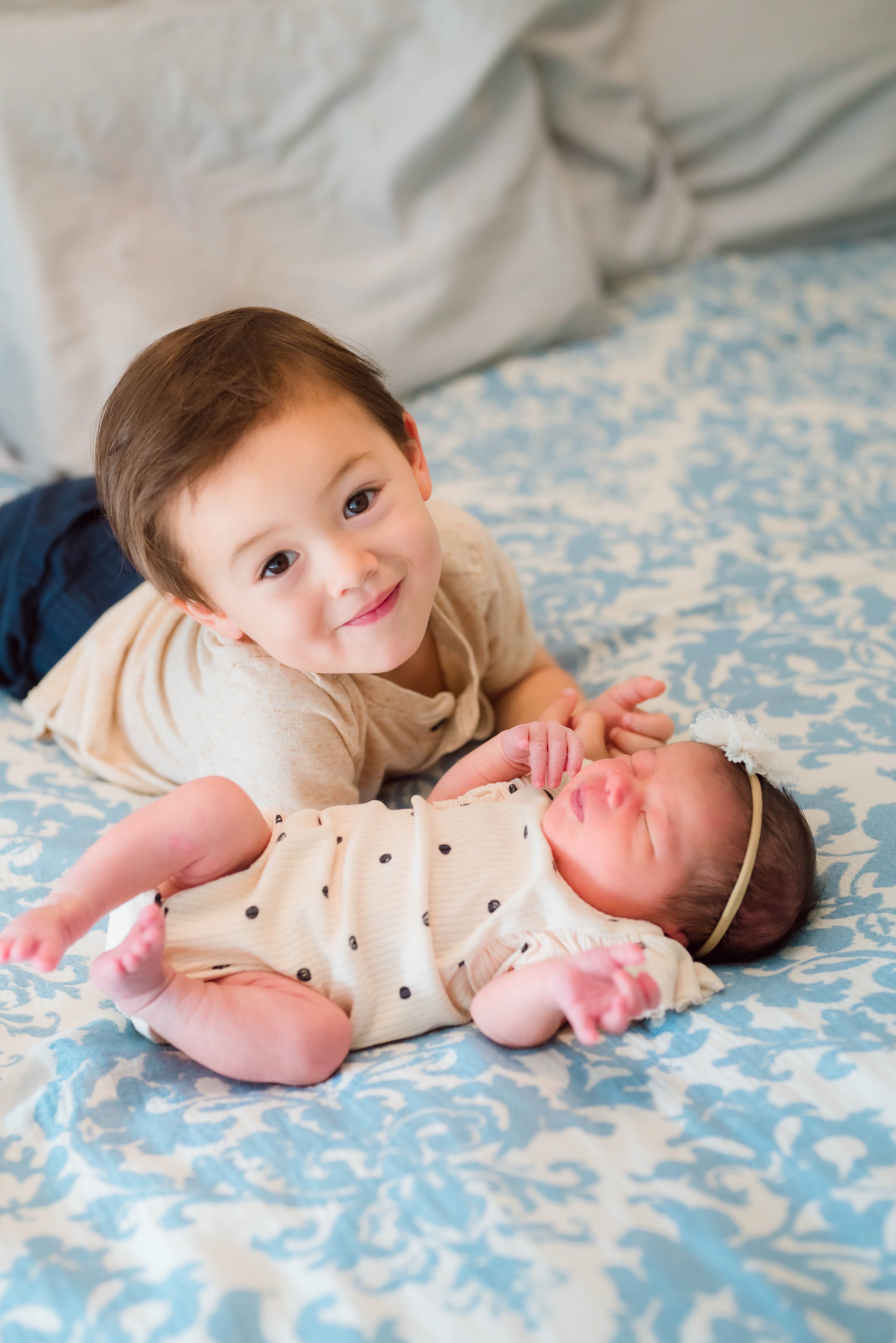 A young boy lying on a bed with a baby girl, both smiling and looking at the camera.