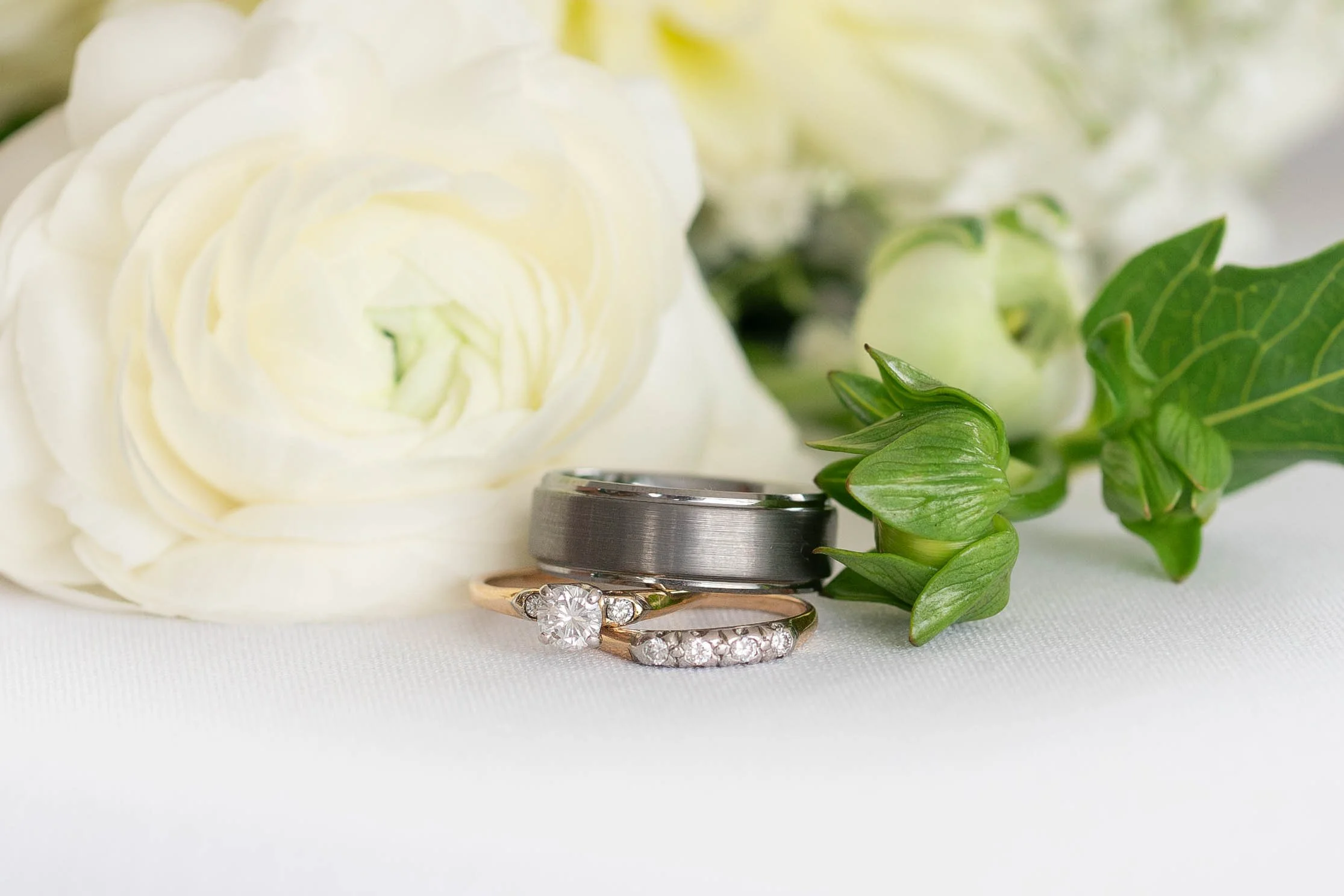 A close-up of wedding rings, one with diamonds, placed on a white surface near a white rose and green leaves.