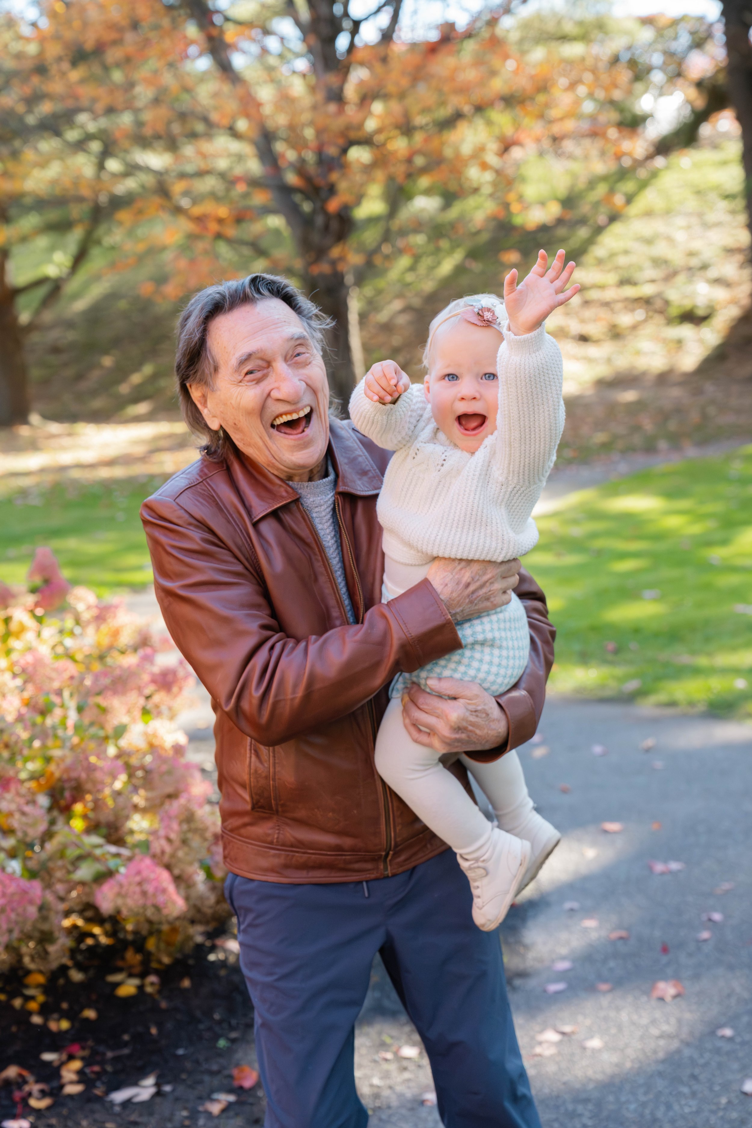 An elderly man in a brown leather jacket smiling and holding a young girl with blonde hair and blue eyes, dressed in a white sweater and leggings, outdoors in a park during autumn.