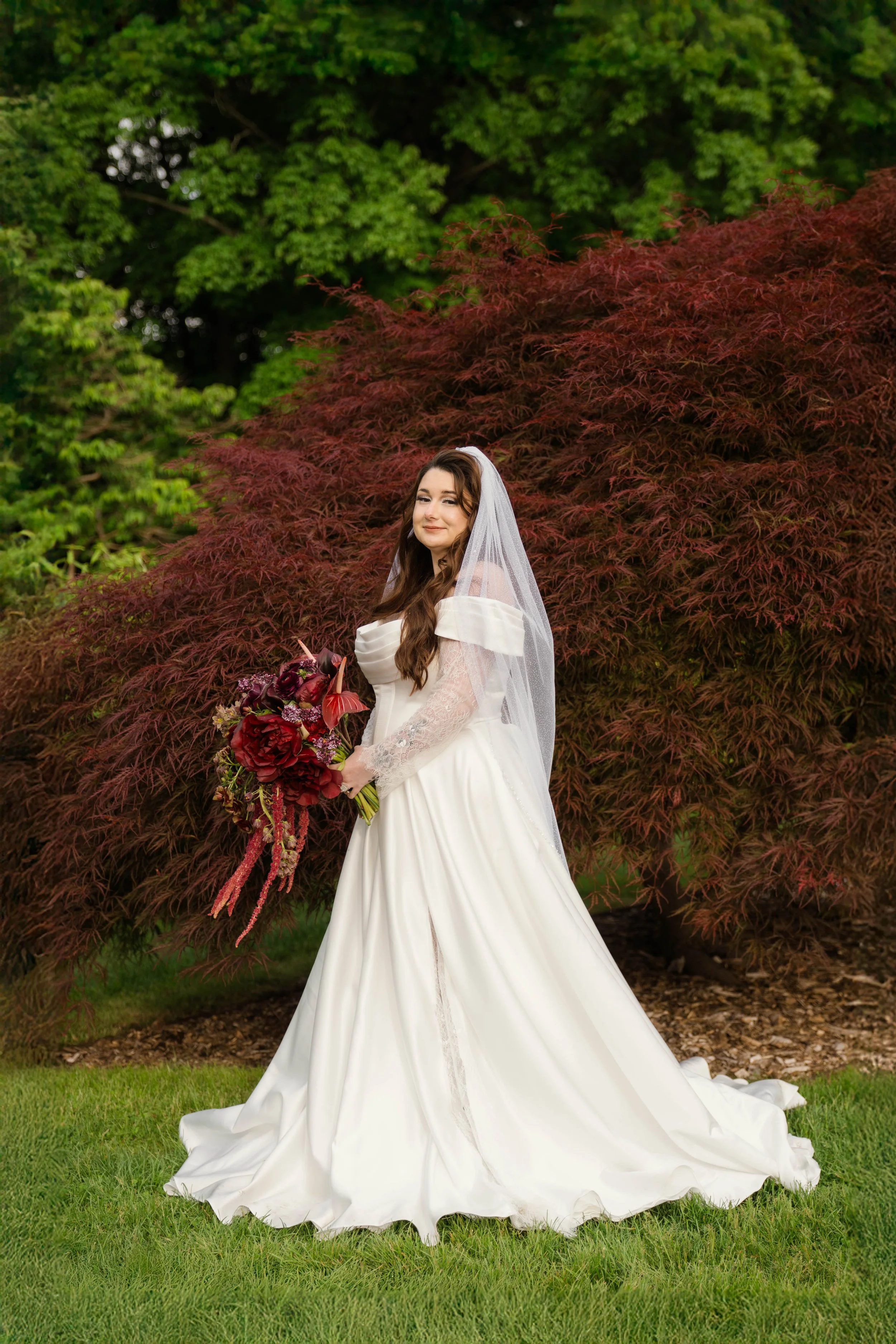 A bride in a white wedding gown with long sleeves, holding a bouquet of burgundy and purple flowers, standing outdoors in front of red and green trees.
